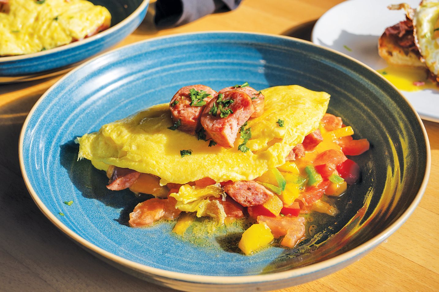 A sausage and vegetable omelet served in a blue bowl, with other breakfast dishes visible in the background.