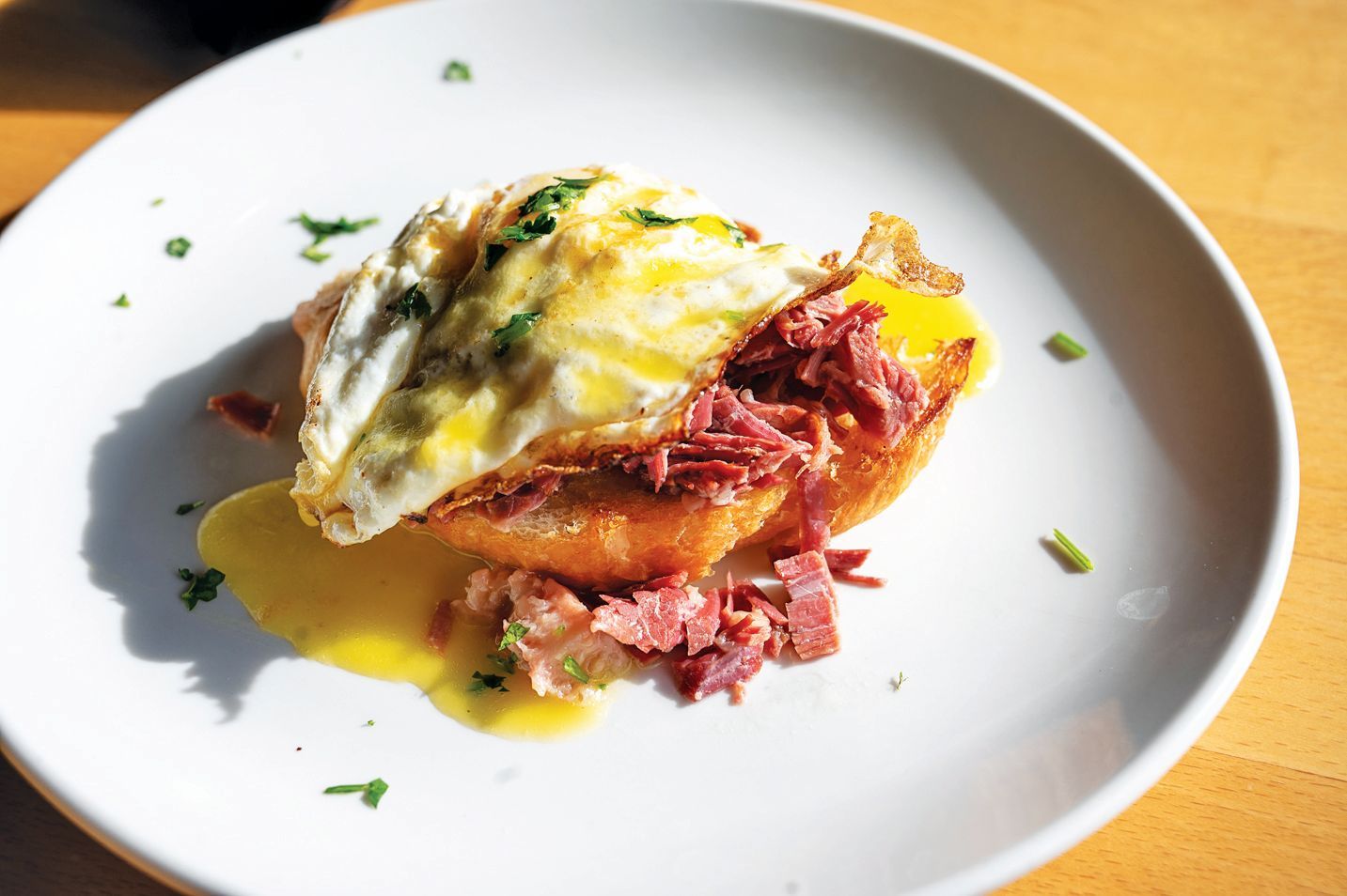 A sunny-side-up egg over corned beef hash and sauce on a white plate, served on a wooden table.
