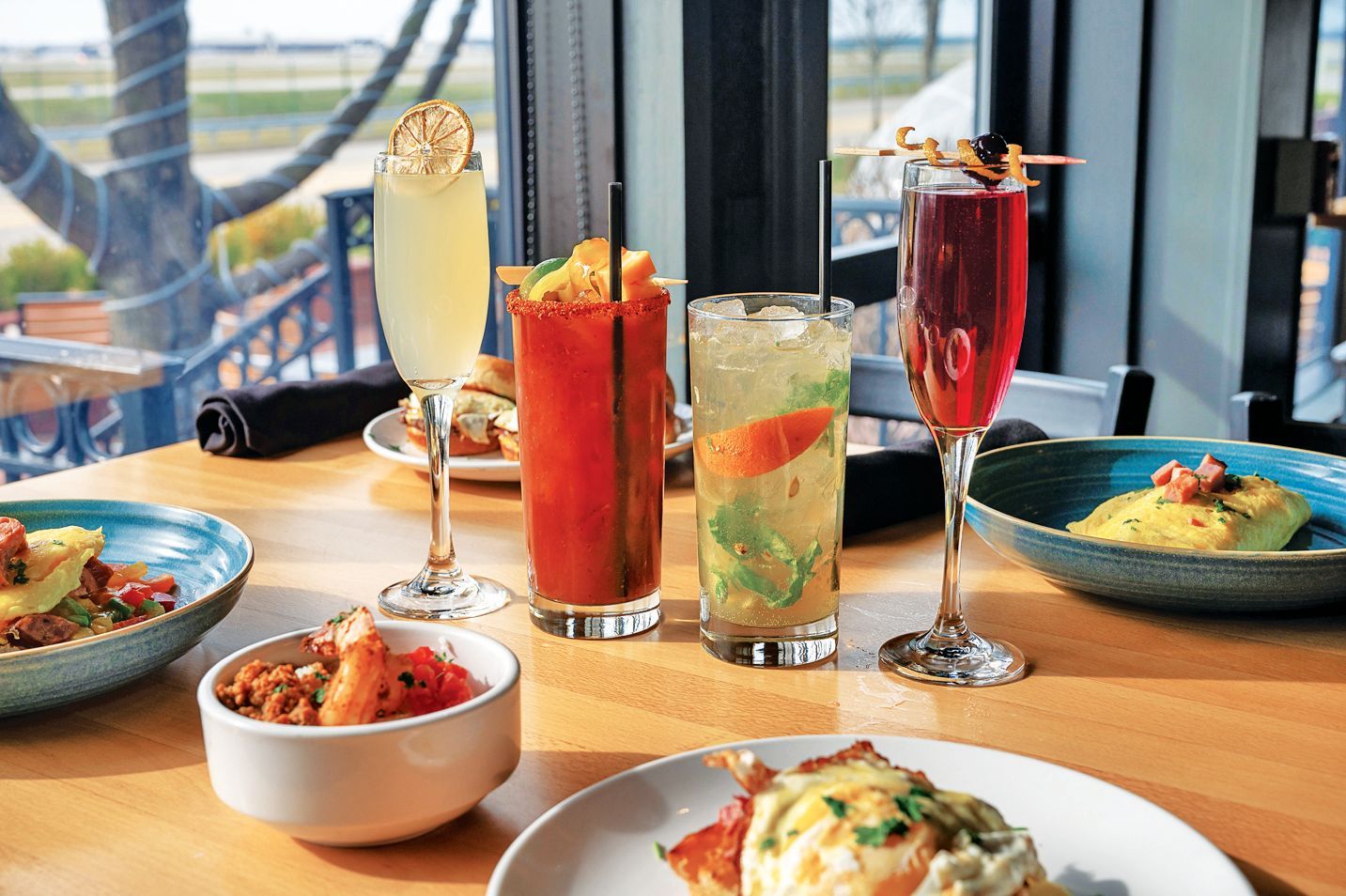 A table at a restaurant with four cocktails, including a Bloody Mary and mimosas, surrounded by plates of brunch food.