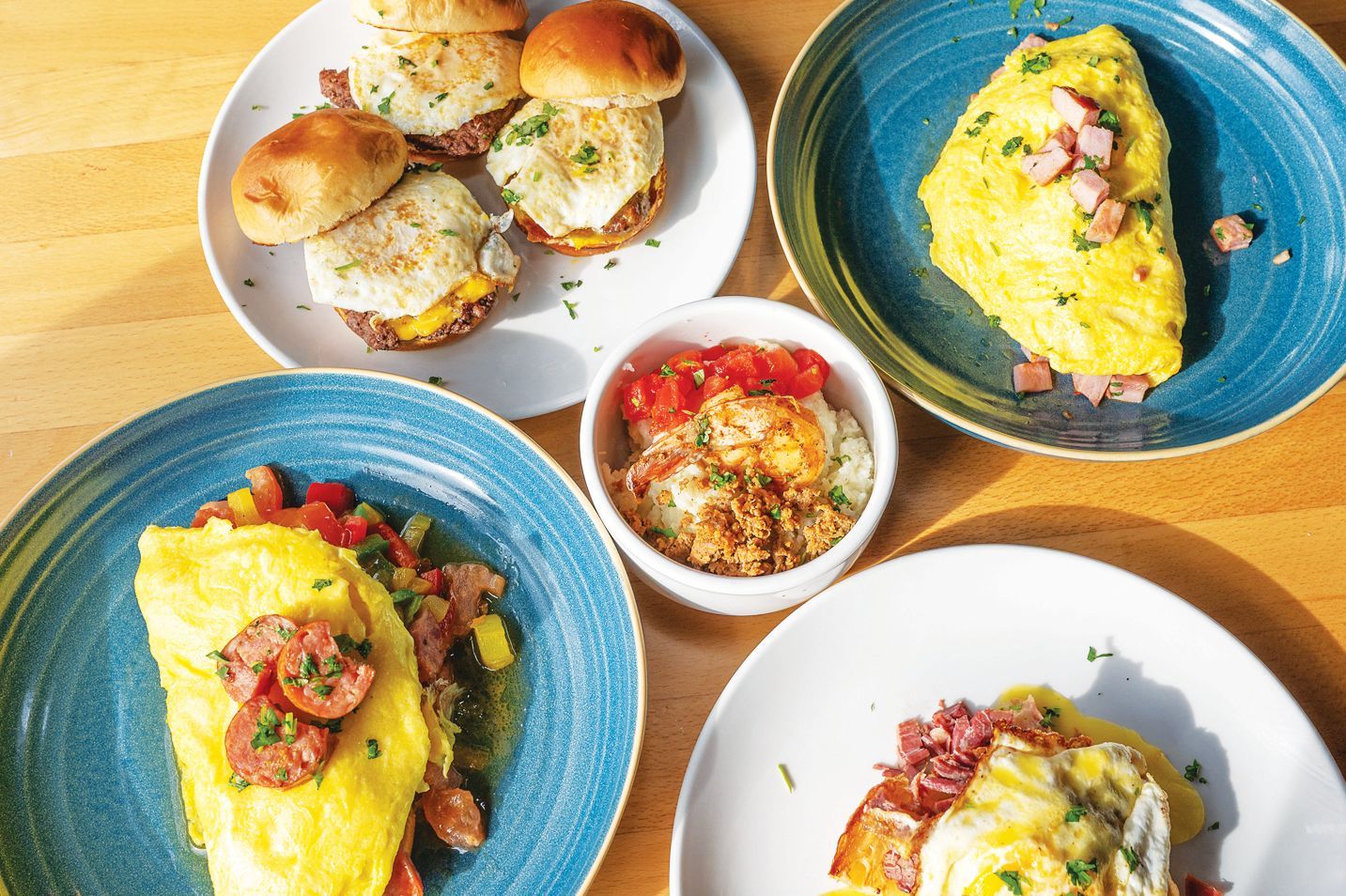 Top-down view of a wooden table featuring various breakfast dishes, including omelets, breakfast sliders, and a side bowl.