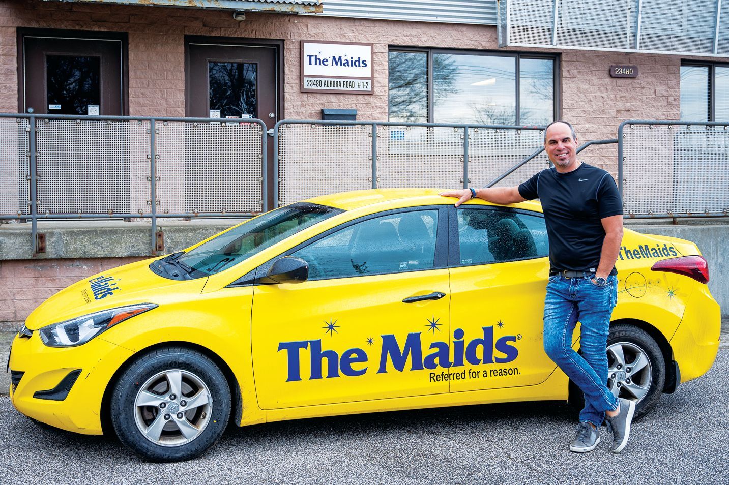 A smiling person leans against a bright yellow company car with 