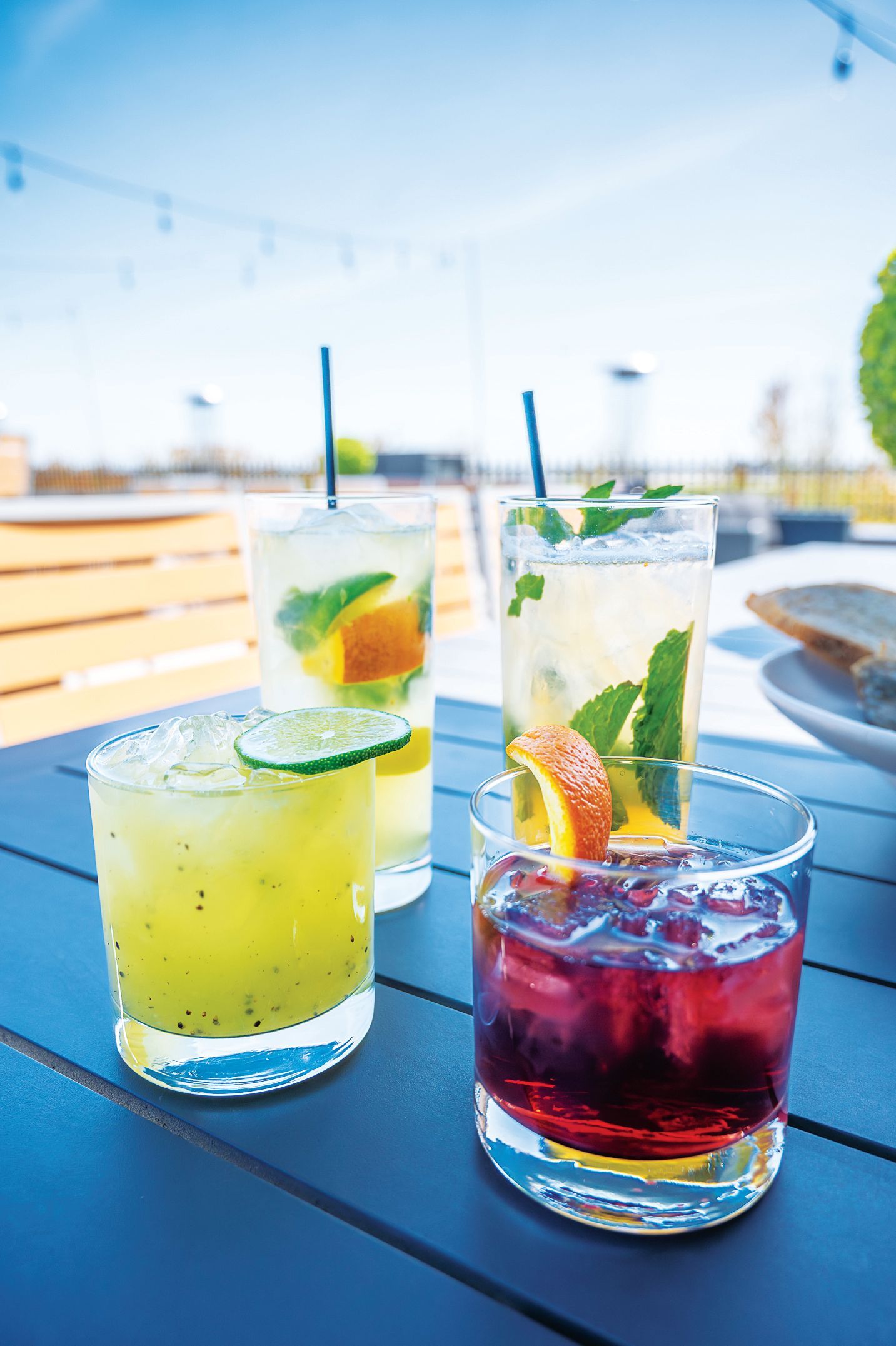 Four colorful cocktails with fruit and herb garnishes arranged on a blue outdoor table on a sunny day.