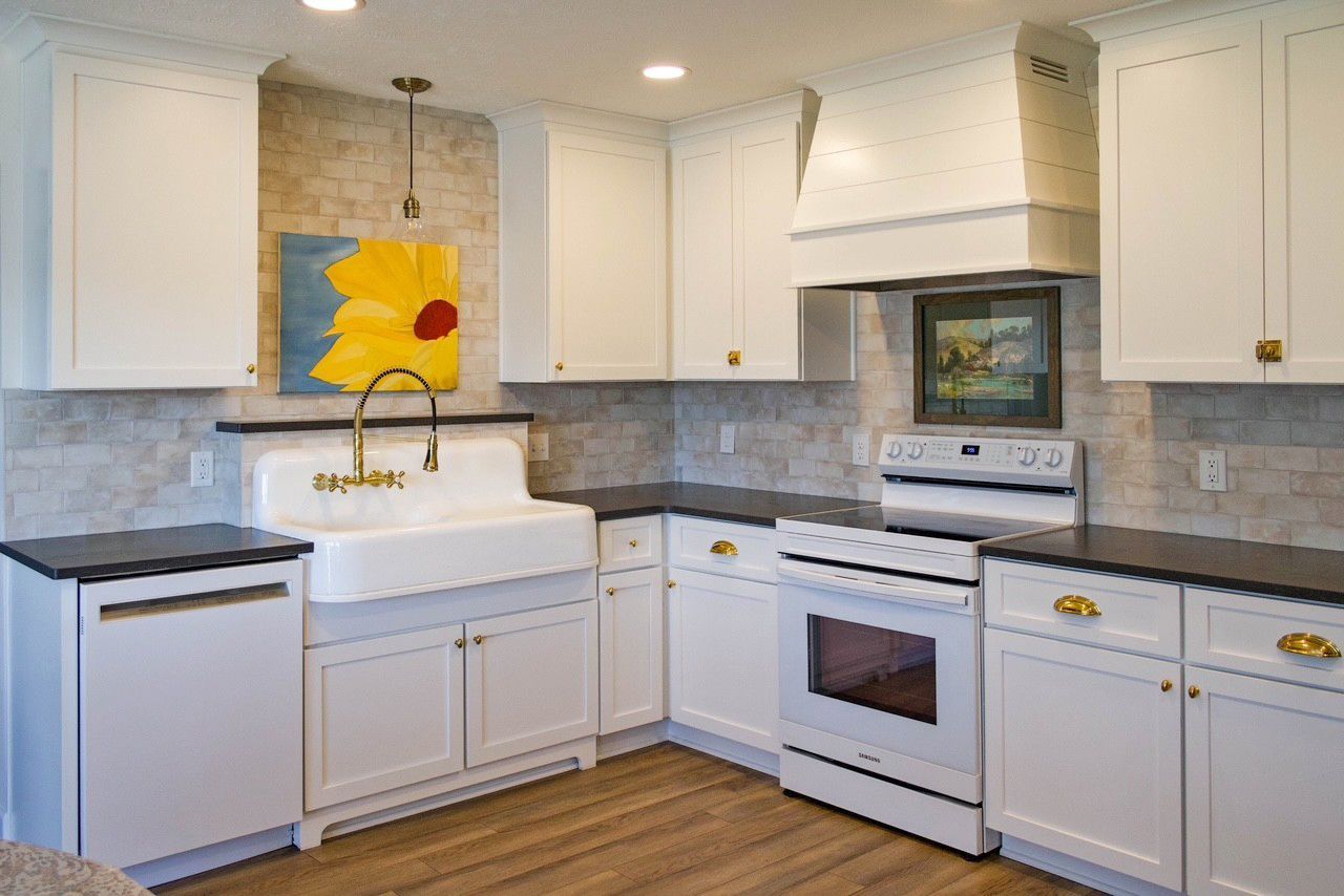 A kitchen with white cabinets, a farmhouse sink, dark countertops, a white oven, and a yellow flower painting on the wall.
