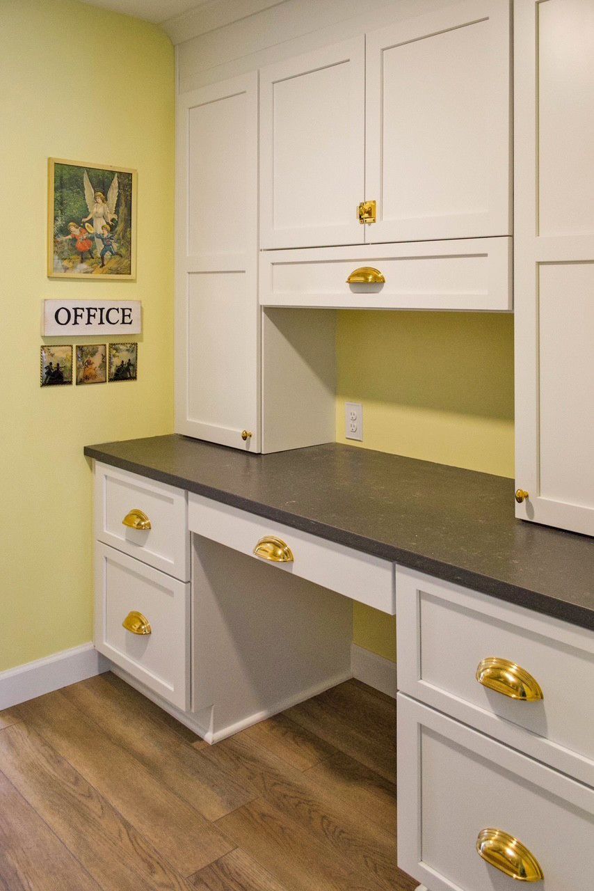 A white built-in desk with dark counters, brass hardware, and yellow walls in a home office setting.
