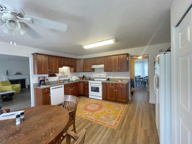 A kitchen with brown wooden cabinets, a white stove and dishwasher, and a round wooden table, opening into a living area.