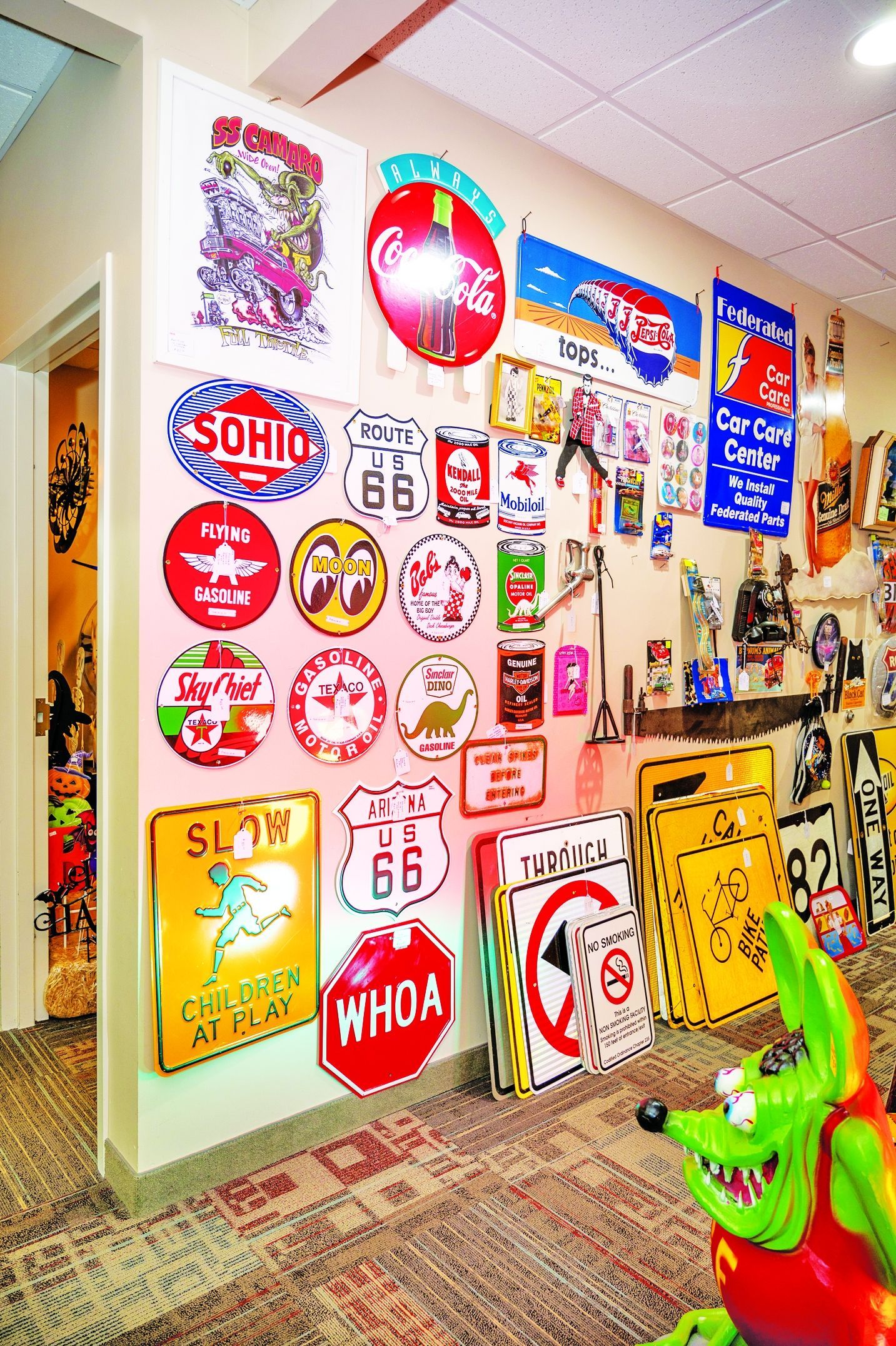 A wall covered in various vintage-style signs, including Coca-Cola, Route 66, and Sohio, next to a lime green character statue.