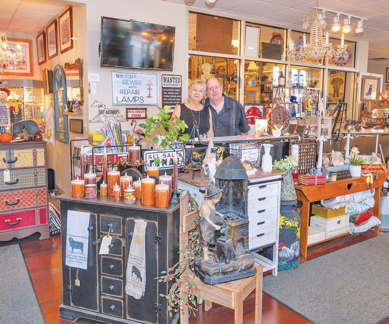 A couple stands behind the counter of a cluttered antique shop filled with home decor, furniture, and vintage items.
