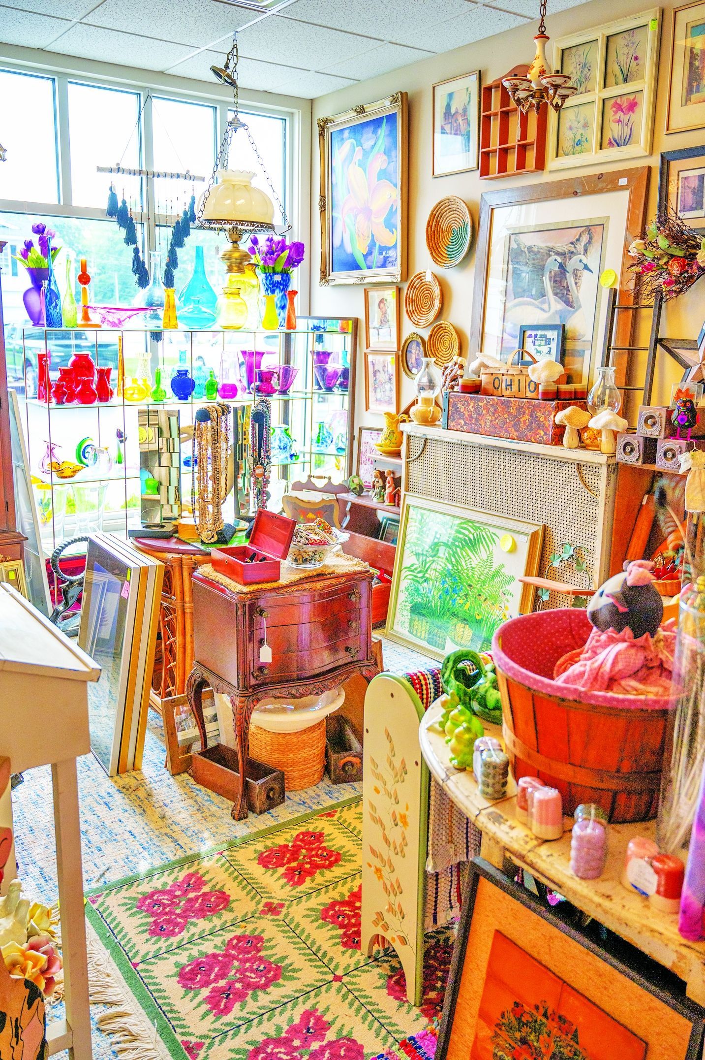 A cluttered, colorful vintage shop interior filled with glassware, paintings, decor, and a patterned rug on the floor.