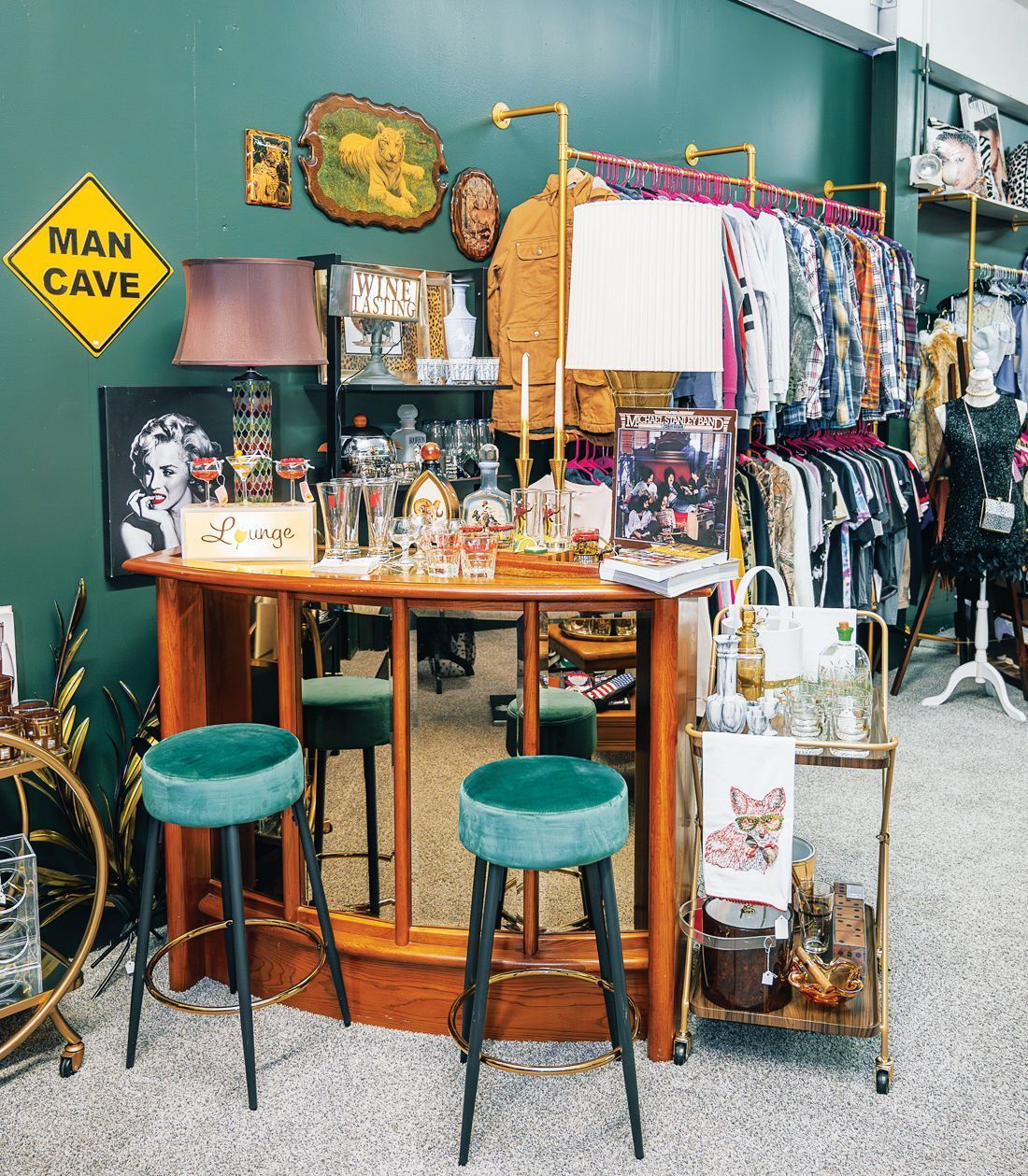 A vintage-style wooden bar with two teal stools, topped with lamps and glassware, set against a dark green wall.