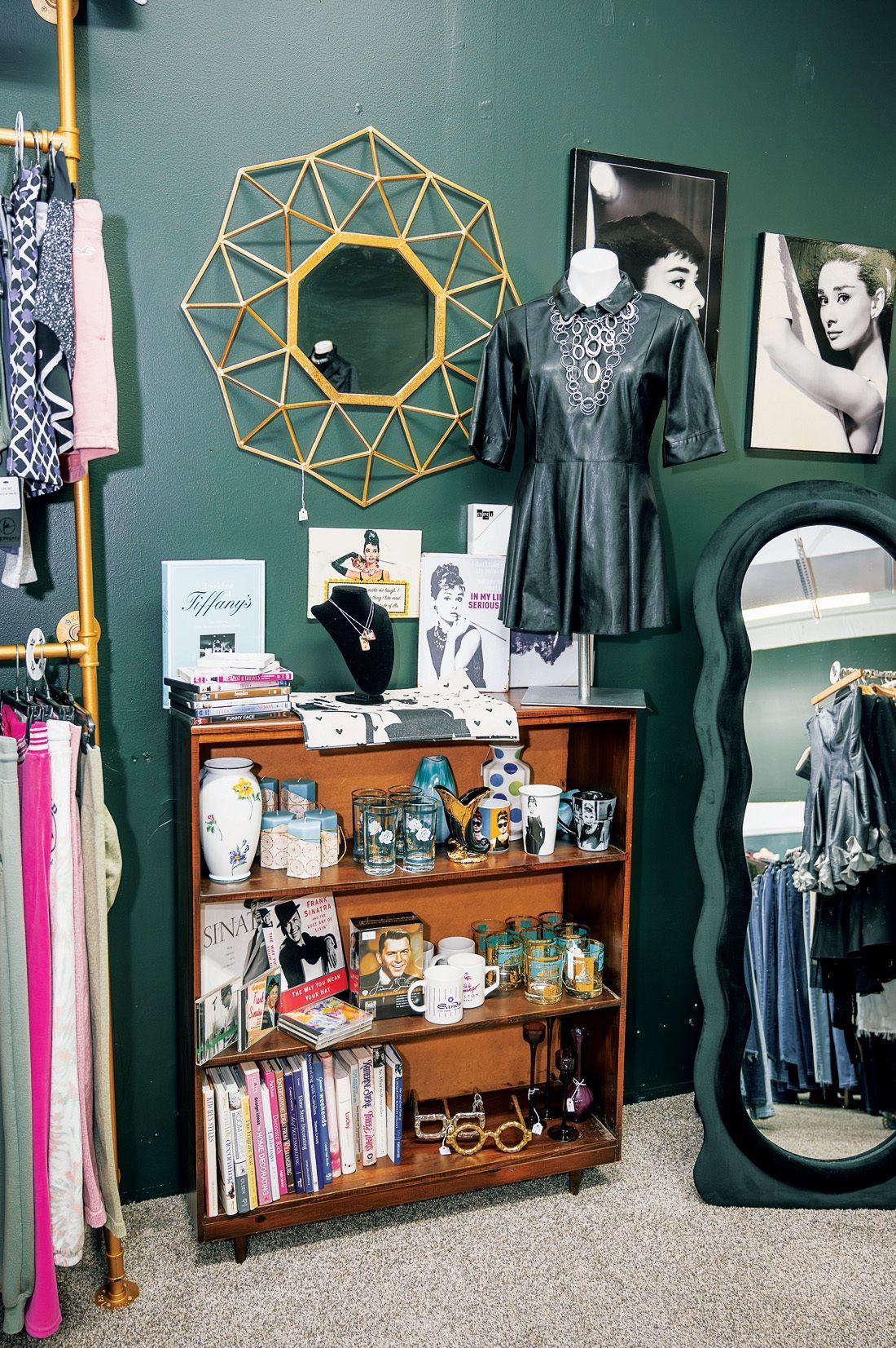 A display of clothing, decor, and accessories in a boutique with a deep green wall, wooden shelf, and oval mirror.