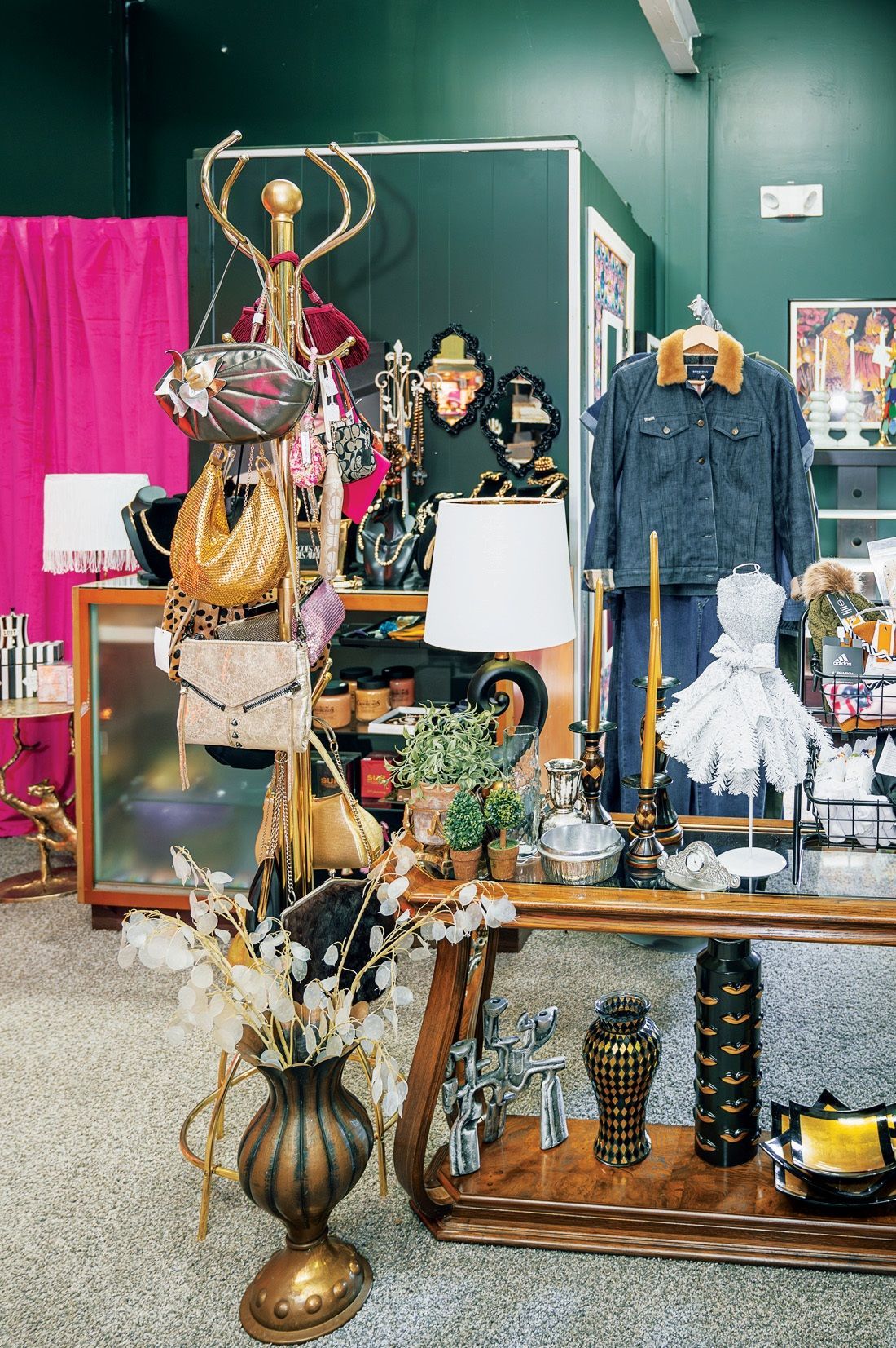 A shop display with a gold coat rack holding purses, a denim jacket on a mannequin, a lamp, and decor on a wooden shelf.