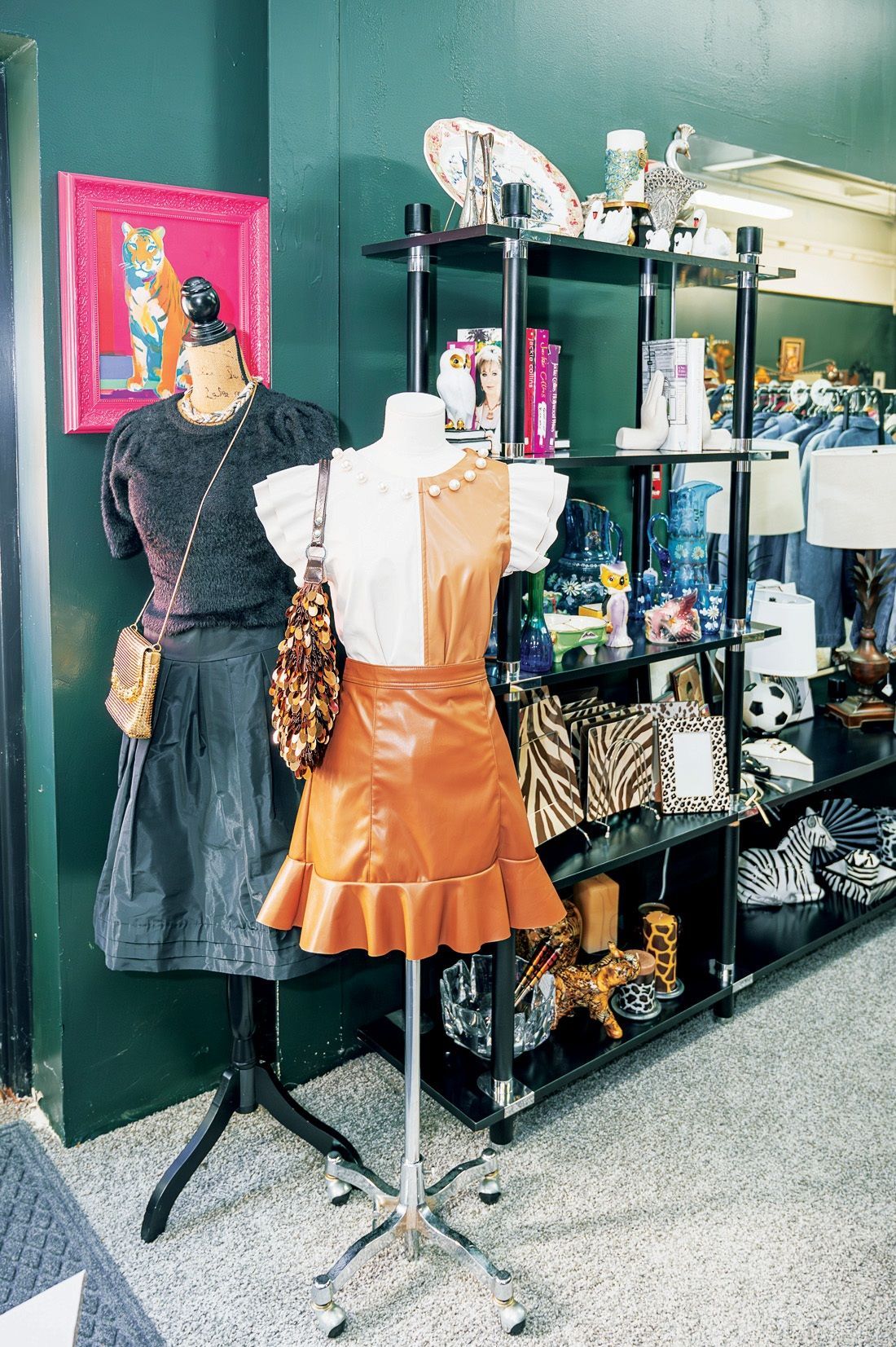 Two mannequins display clothing in a shop with a green wall, a framed picture, and shelves of decorative items.
