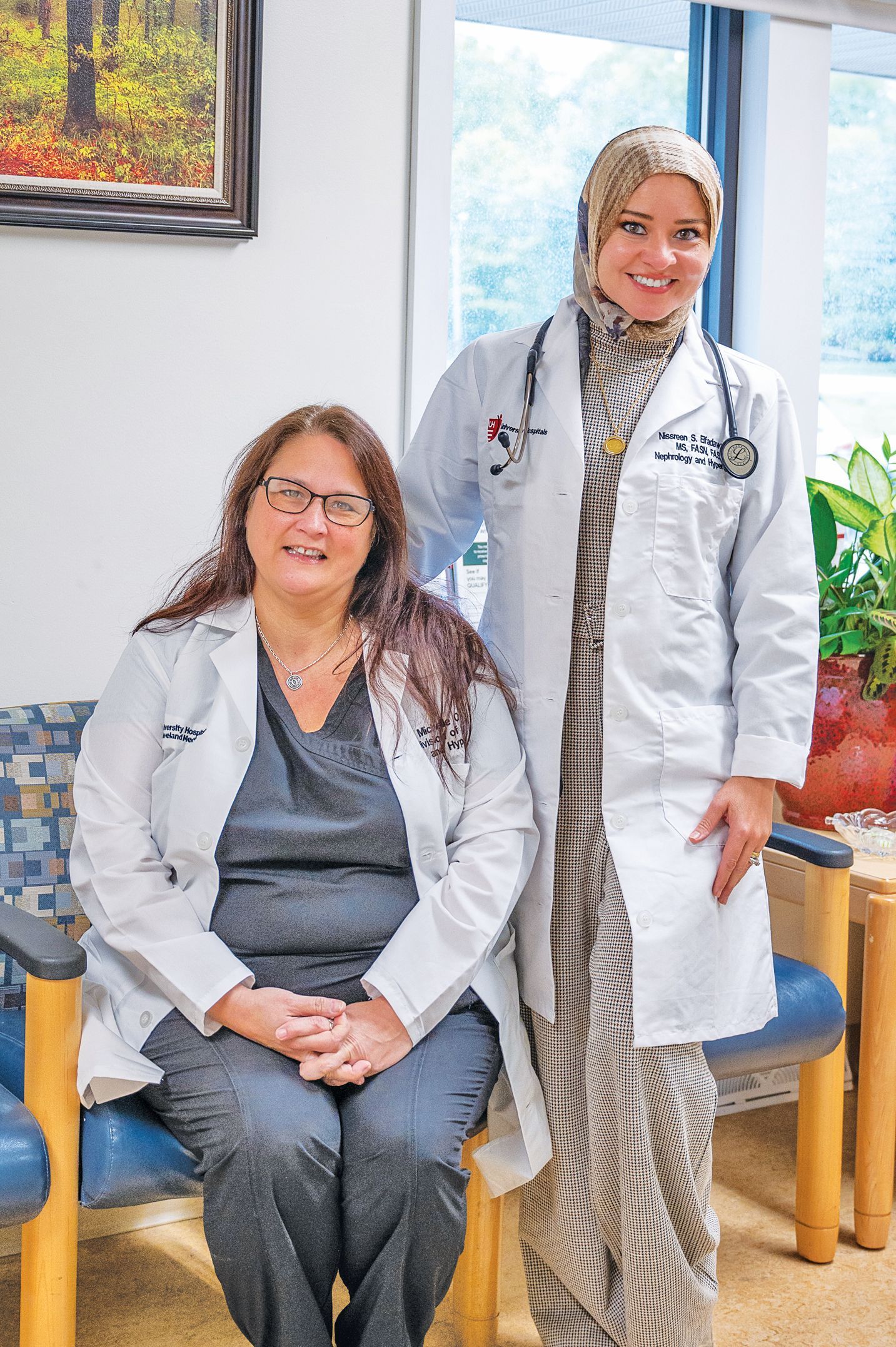 Two professionals in white lab coats and stethoscopes pose in an office setting.