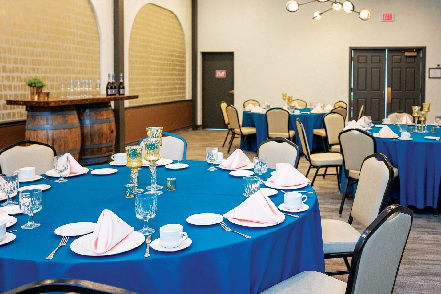 A dining room with round tables covered in blue cloths, set with white plates, napkins, and chairs for an event.