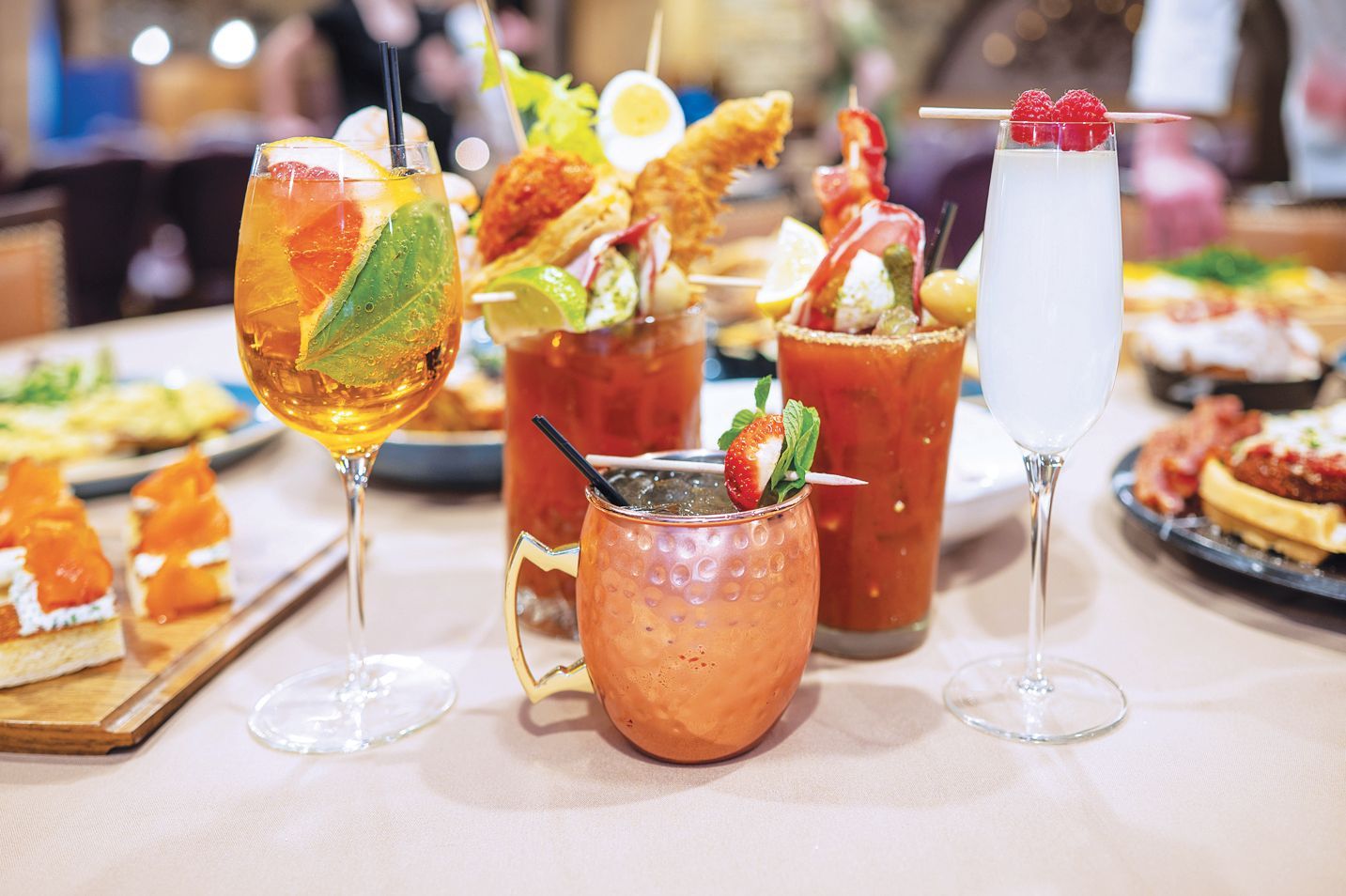 A table setting with four cocktails, including two elaborate Bloody Marys topped with garnishes and a white mimosa.