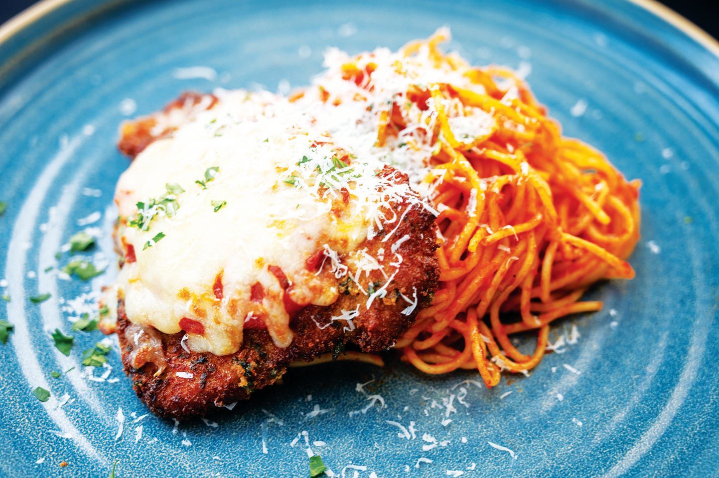 A portion of chicken parmesan topped with melted cheese and red sauce, served with spaghetti on a blue plate.