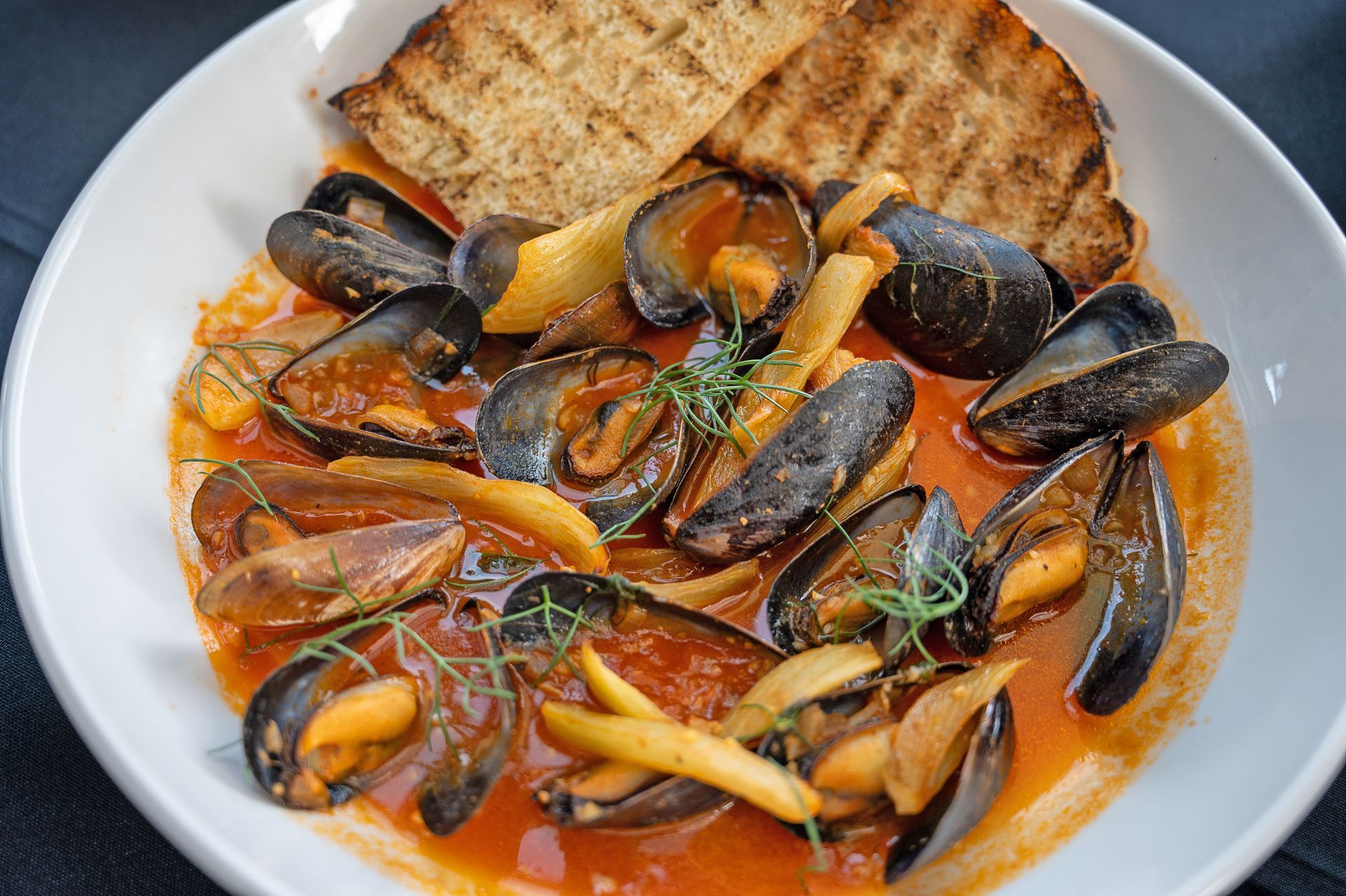 A bowl of mussels in a savory red tomato sauce, garnished with herbs and served with two pieces of toasted bread.