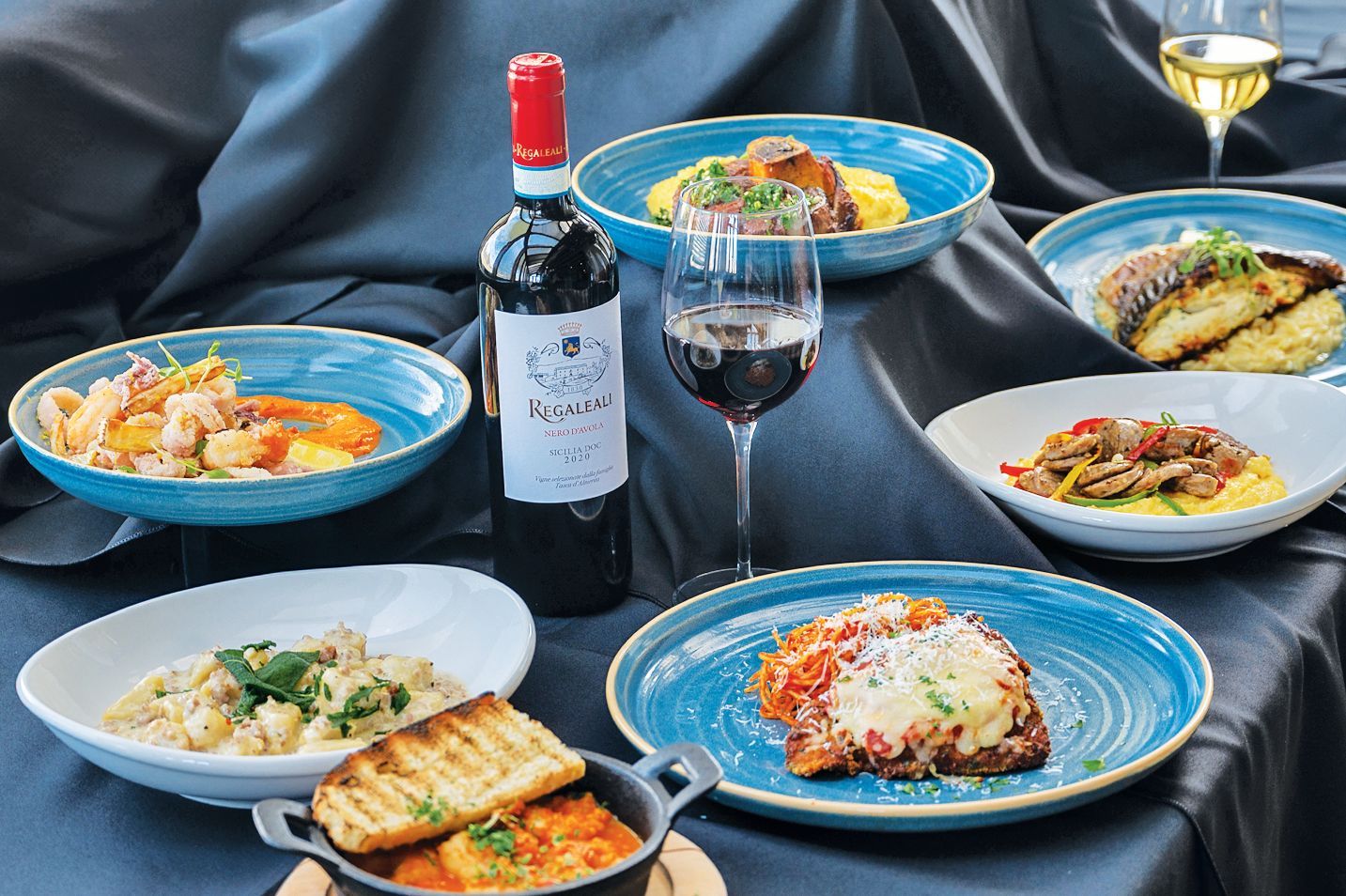 A variety of gourmet dishes served in bowls and on plates, arranged on a black tablecloth with wine.
