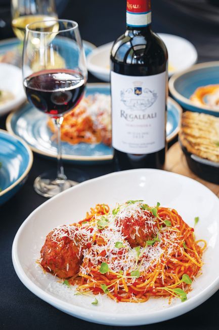 A bowl of spaghetti and meatballs topped with parmesan and herbs next to a glass and bottle of red wine on a table.