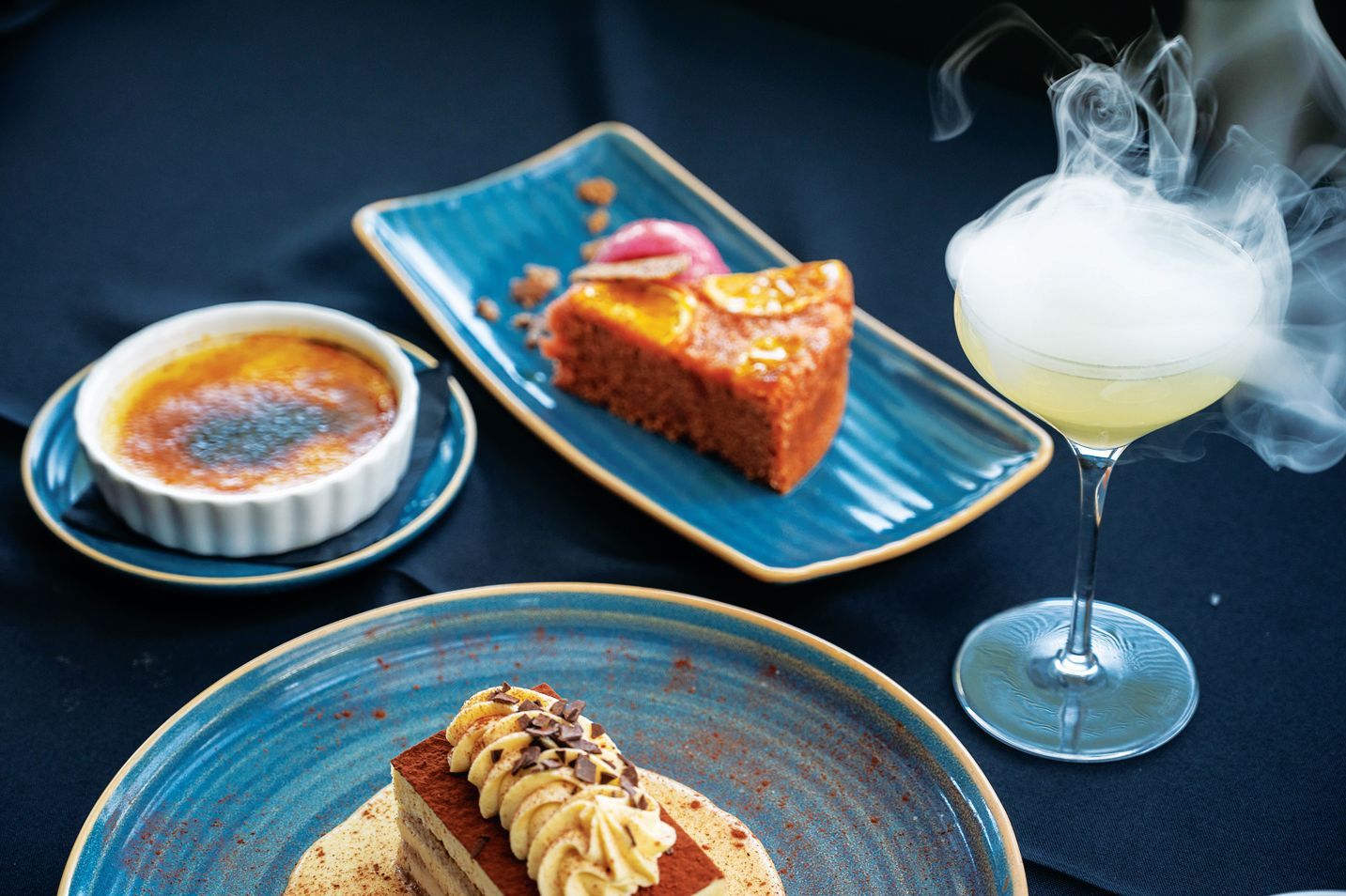 A gourmet dessert spread with a steaming cocktail, crème brûlée, a slice of orange cake, and a decorated pastry.