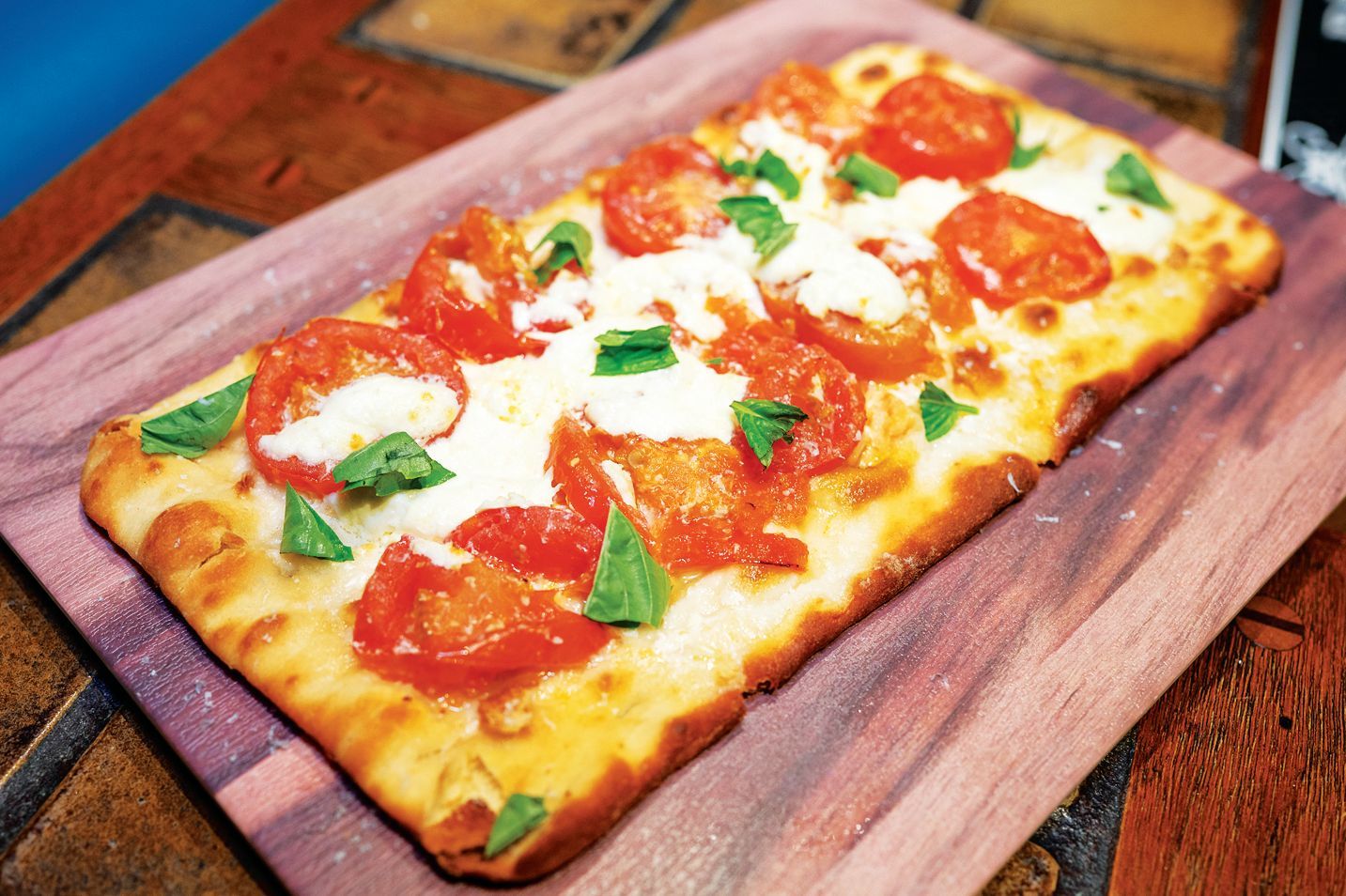 A rectangular flatbread pizza topped with sliced tomatoes, melted mozzarella cheese, and fresh basil on a wooden board.