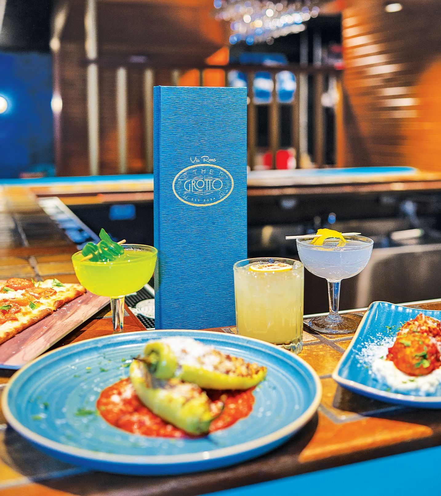 A restaurant bar counter with appetizers, three cocktails, and a blue menu placed in front of a blurred interior.