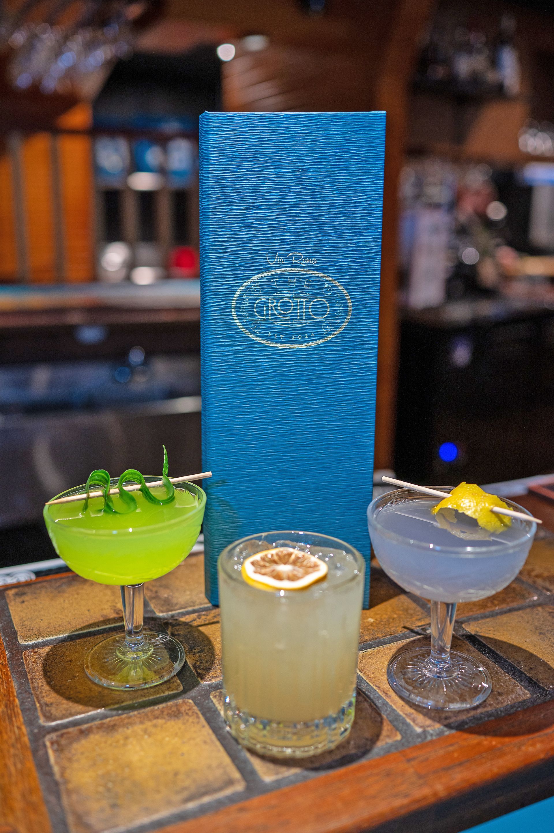 Three cocktails, one bright green, one beige, and one pale blue, stand on a tiled bar in front of a tall blue menu.