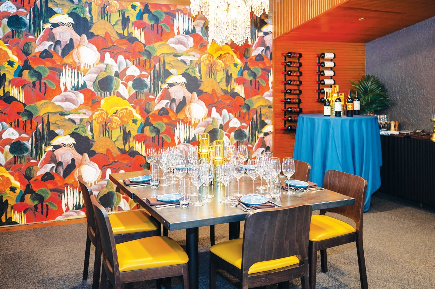 A dining table with place settings, surrounded by chairs with yellow cushions, against a colorful mural wall.
