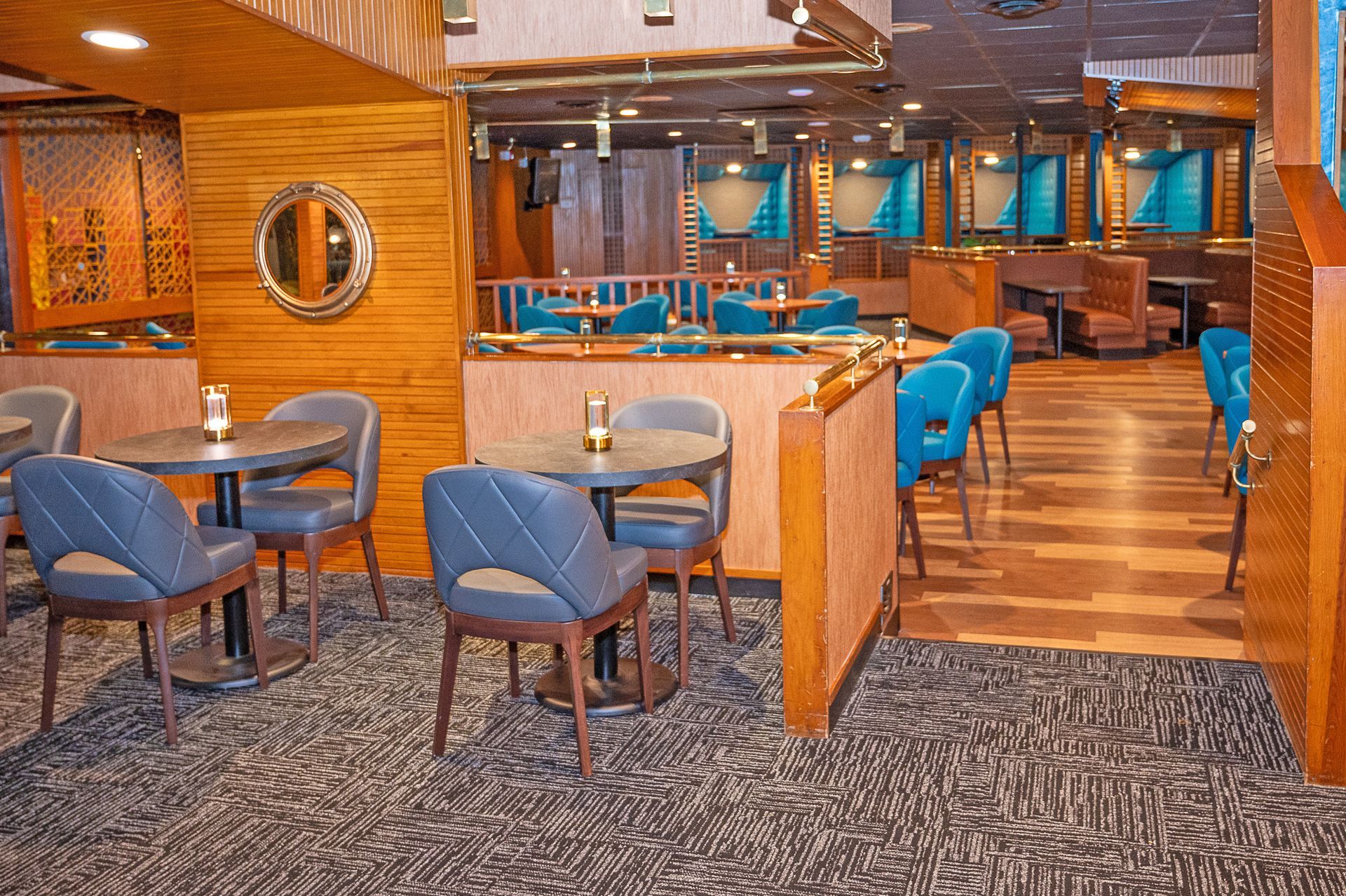 Interior of a restaurant with round tables, blue chairs, wooden accents, and patterned carpeting.