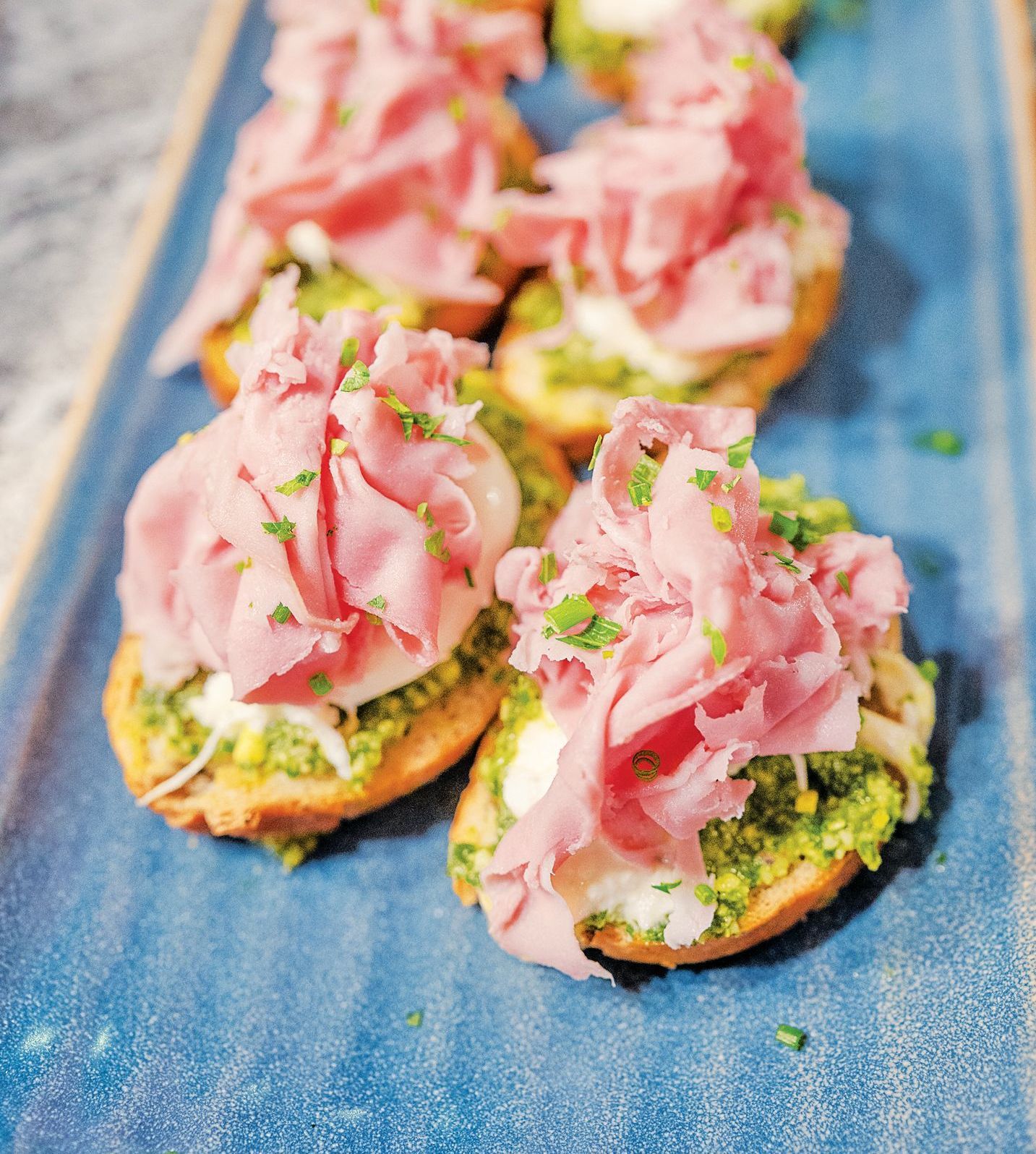 Mortadella topped with pesto and cheese on toasted bread slices, arranged on a textured blue rectangular platter.