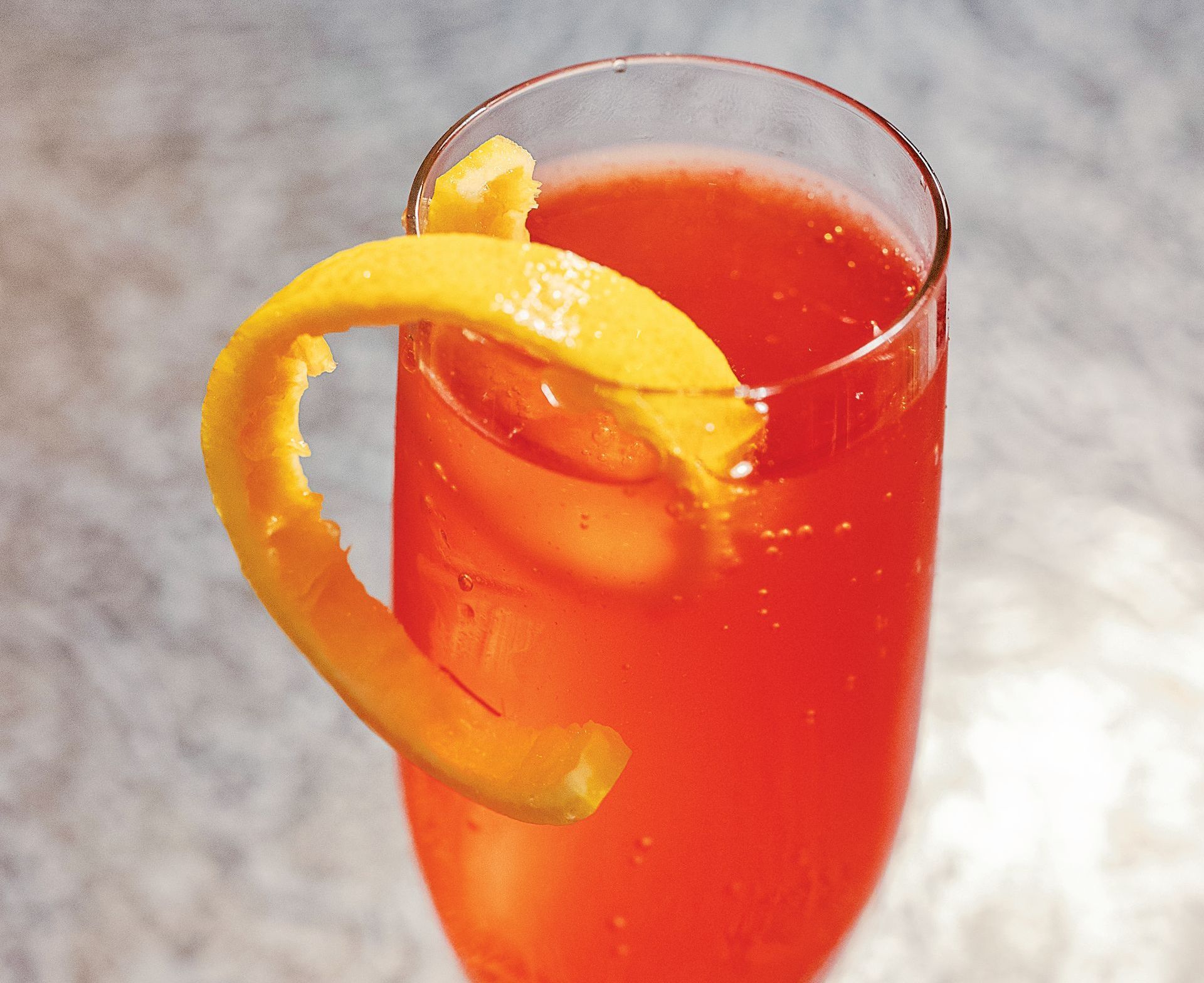 A tall glass filled with a vibrant orange-red cocktail garnished with a thick, curved citrus peel.