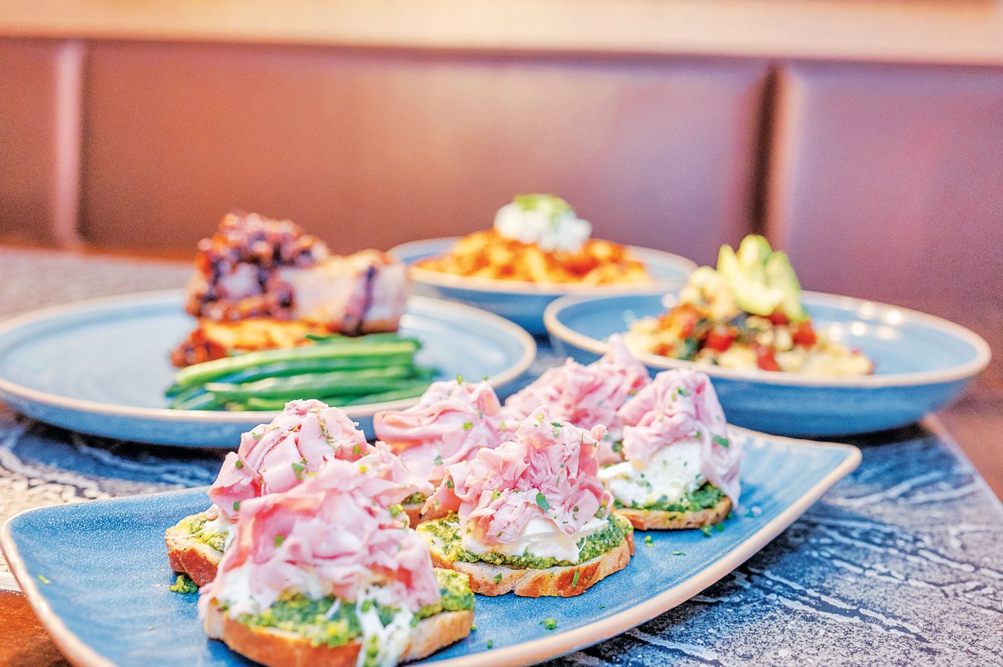 A blue platter with several crostini topped with ham and herbs, set before other plates of food on a table.