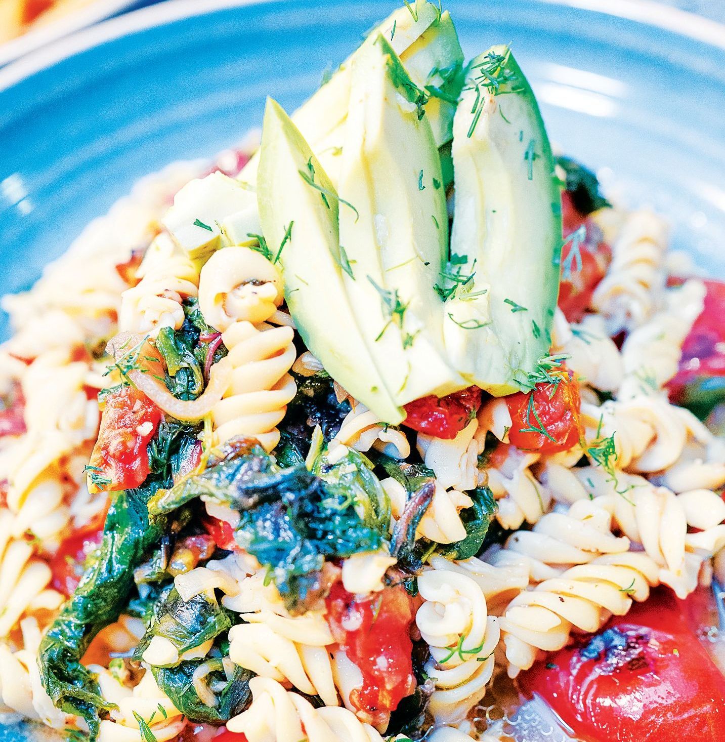 A bowl of rotini pasta tossed with spinach, roasted tomatoes, and fresh dill, topped with sliced avocado.