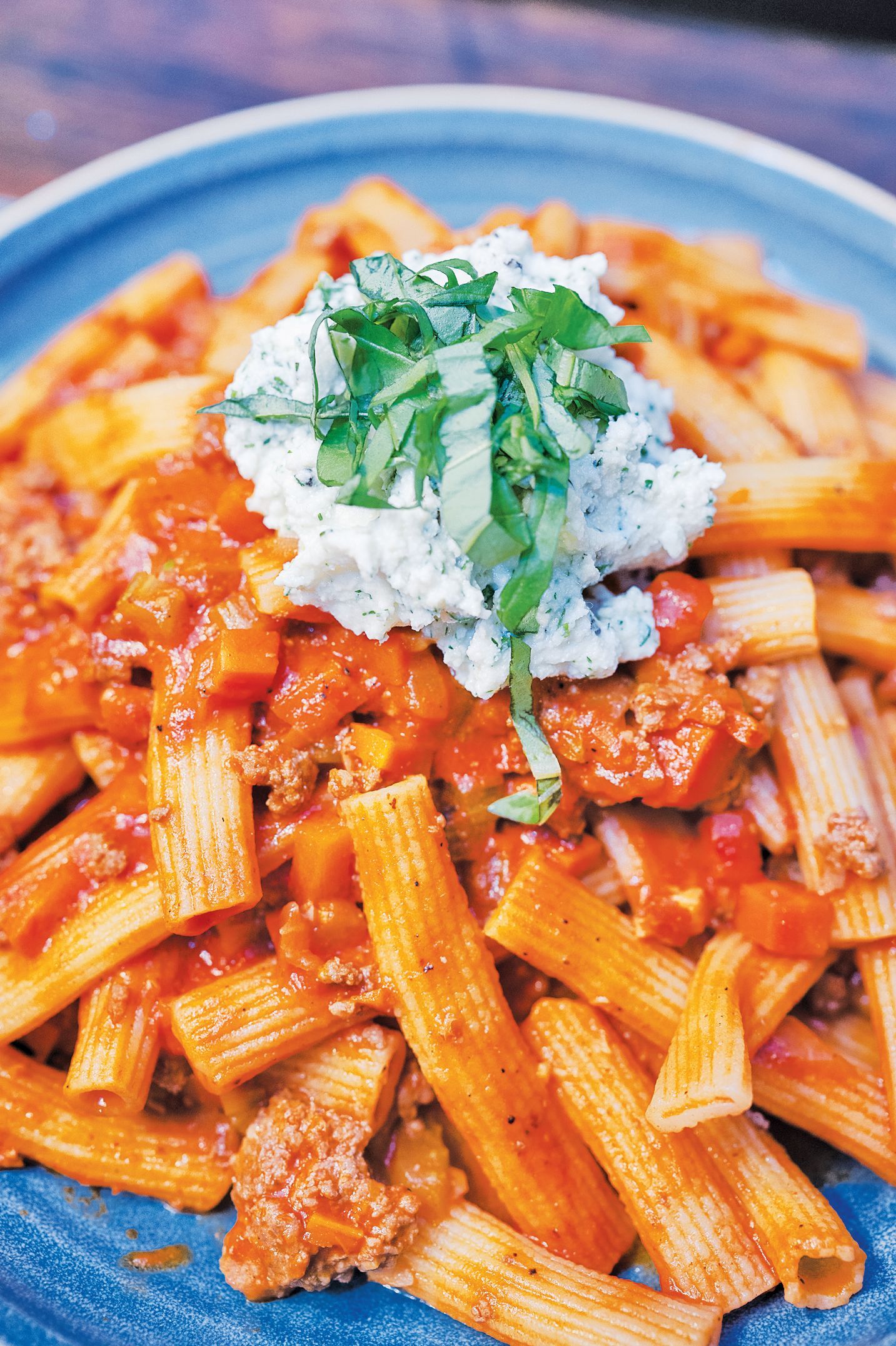 Penne pasta in a meat-based red sauce, topped with a scoop of ricotta cheese and chopped fresh basil on a blue plate.