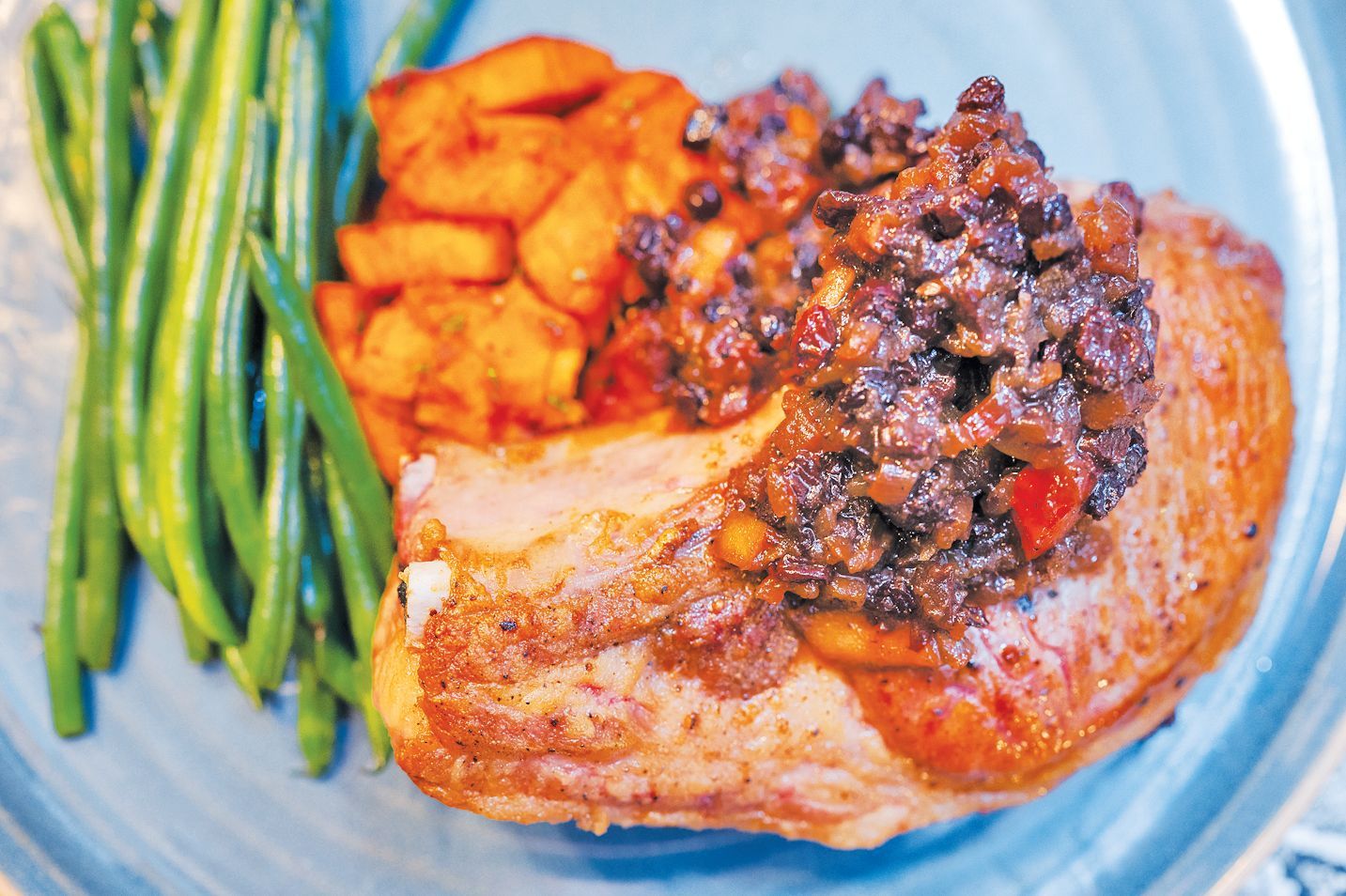 A pork chop topped with a dark bean salsa, served with roasted sweet potatoes and green beans on a blue plate.