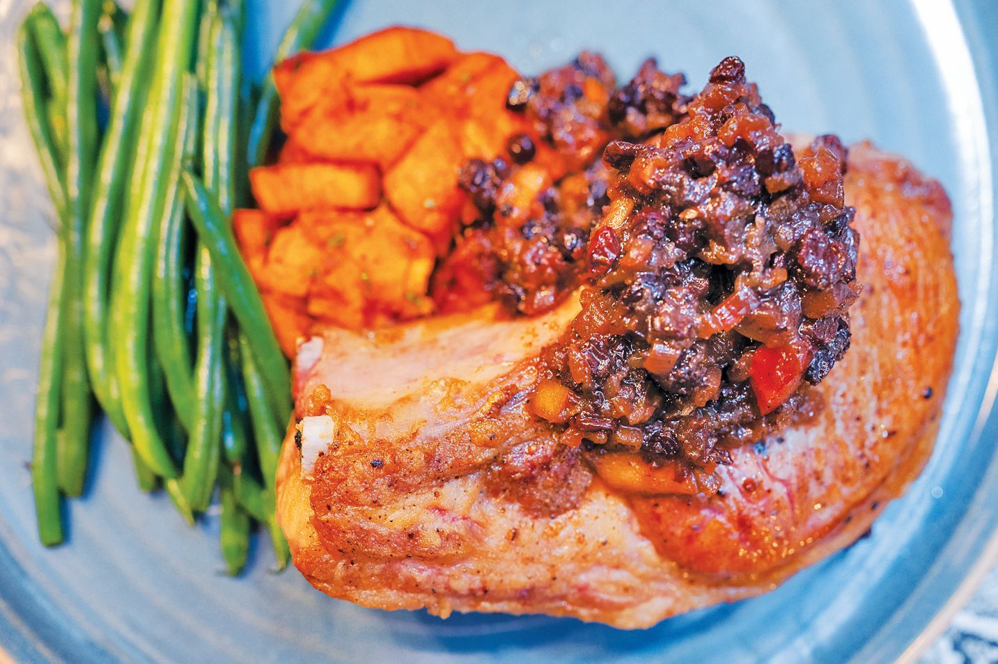 A plate featuring a cooked pork chop topped with a dark tapenade, roasted sweet potatoes, and a side of green beans.
