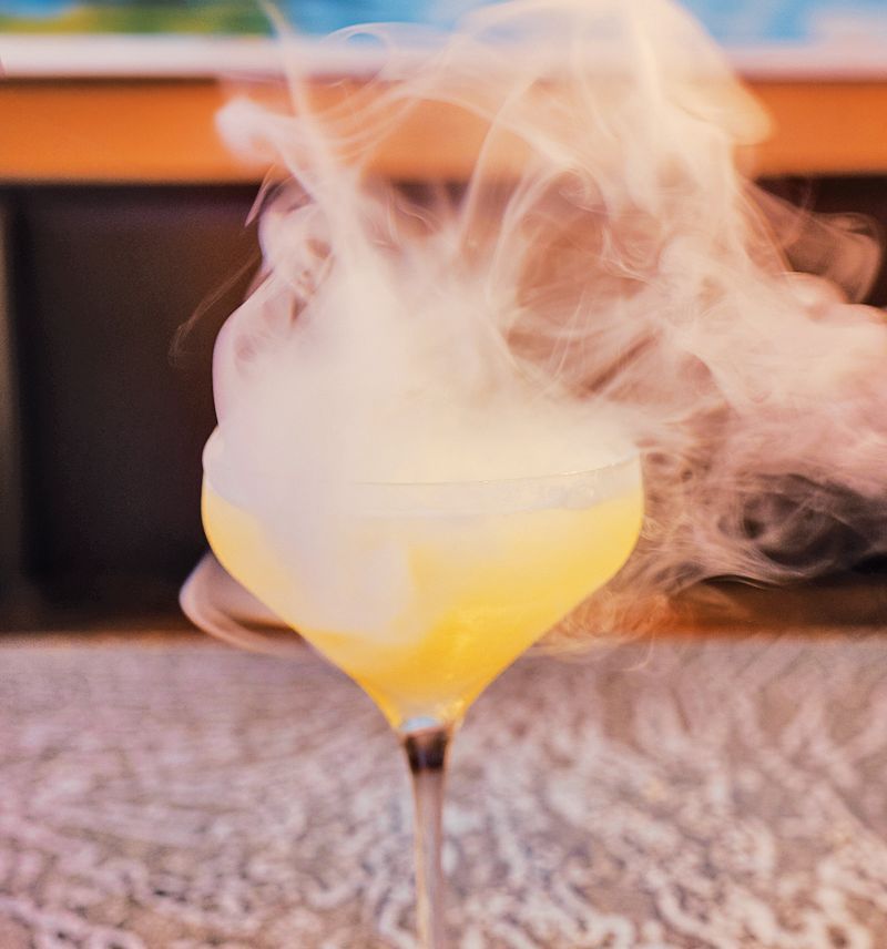 A yellow cocktail in a stemmed glass producing a dramatic, swirling plume of white dry ice smoke.
