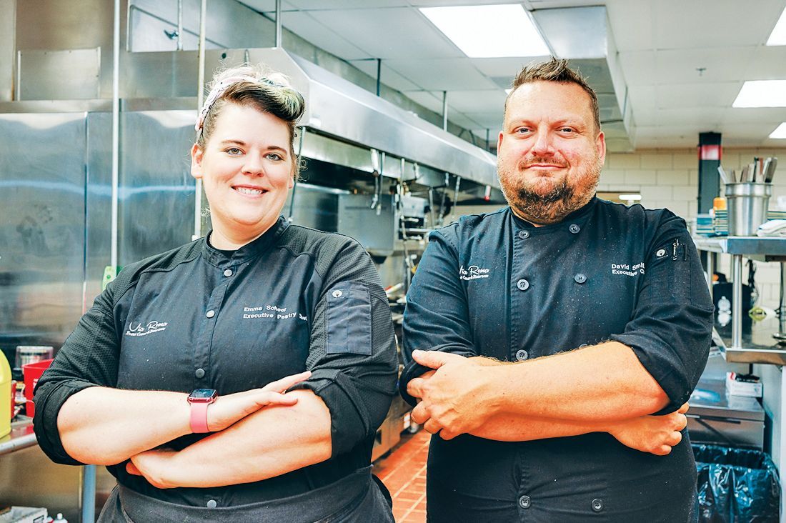 Two people in black chef uniforms with their arms crossed, smiling, standing in a commercial kitchen.