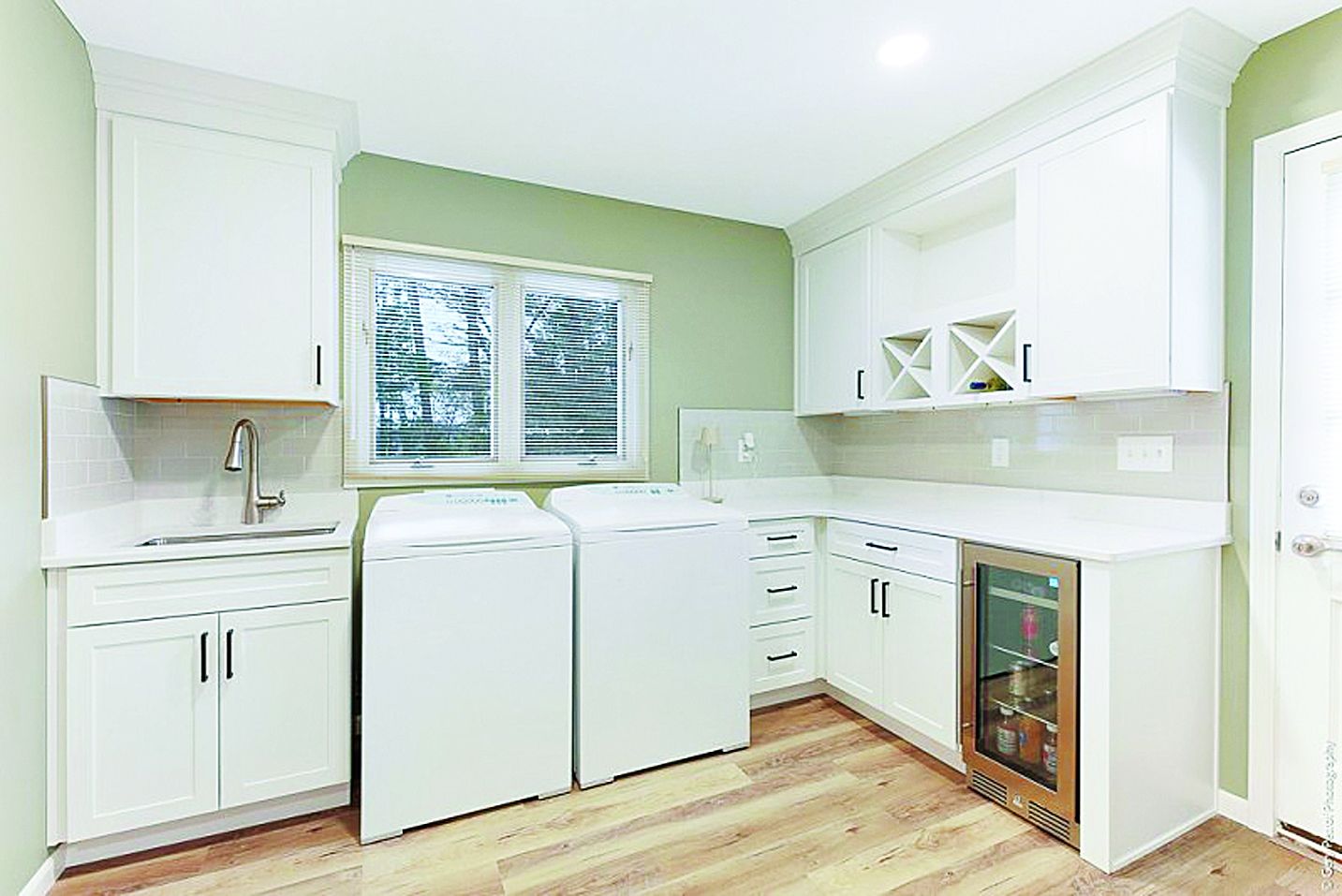 A laundry room with white cabinets, a sink, a washer and dryer, and a built-in wine cooler against sage green walls.