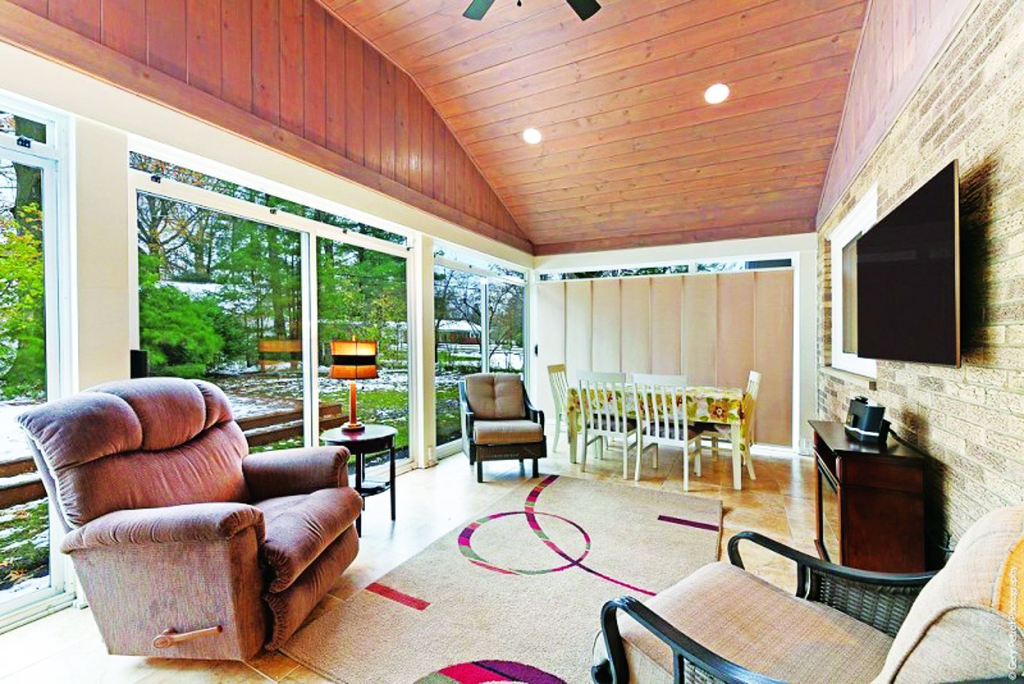 A sunroom with a vaulted wood ceiling, a large armchair, a dining set, and a wall-mounted television.