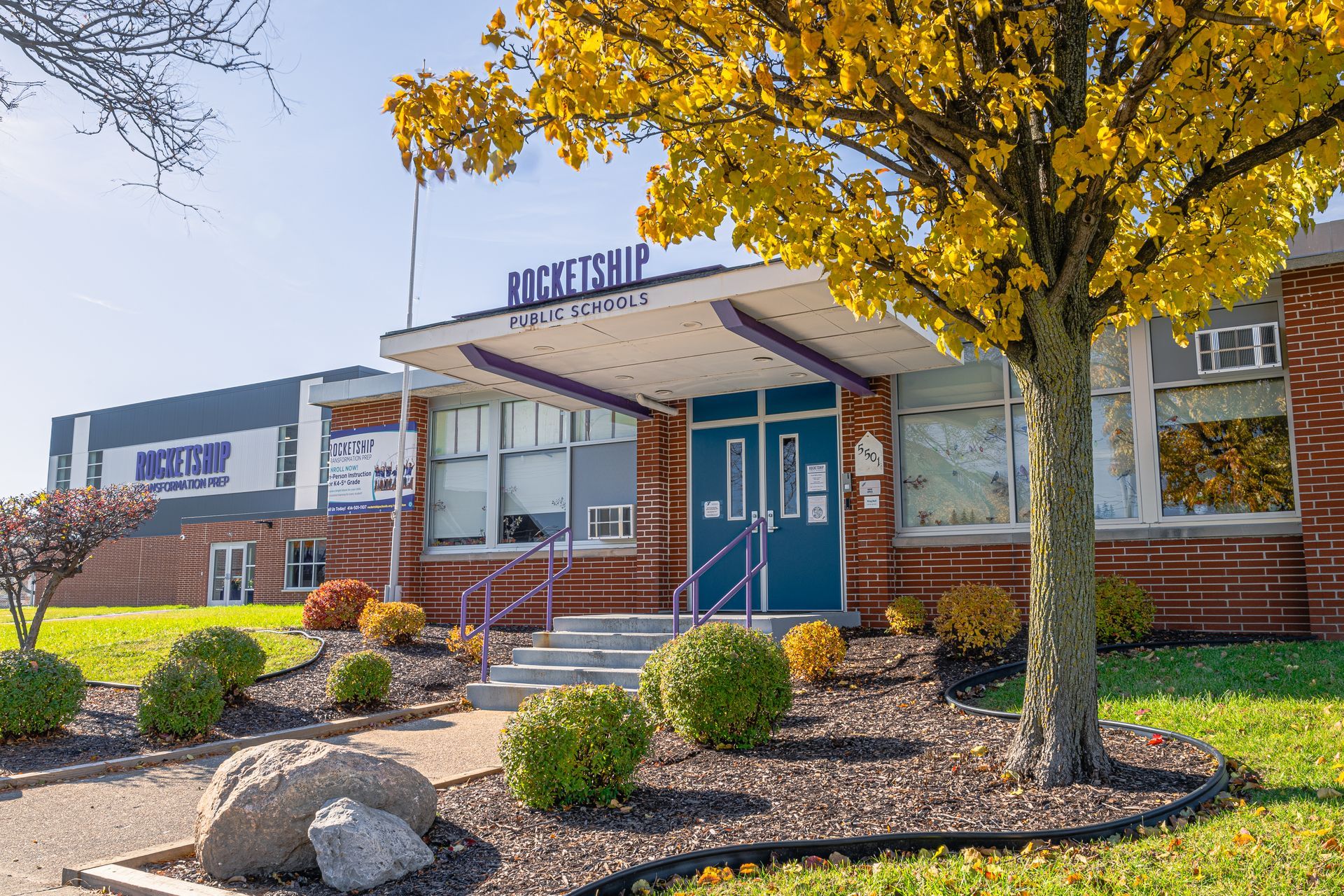 Front entrance of a brick building with blue doors, autumn trees, and a landscaped walkway