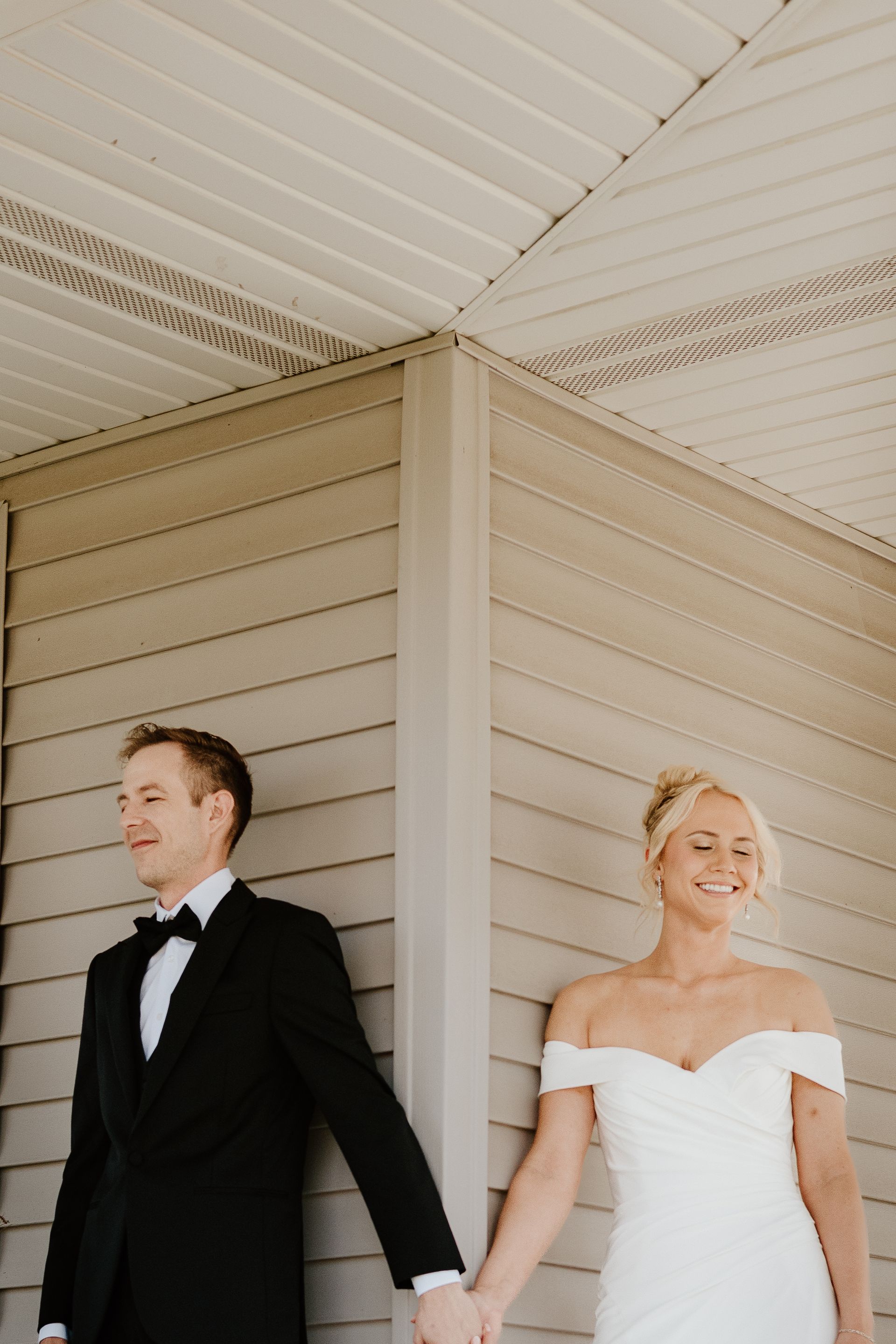 Couple in formal wear standing under a porch, holding hands at a corner of the house