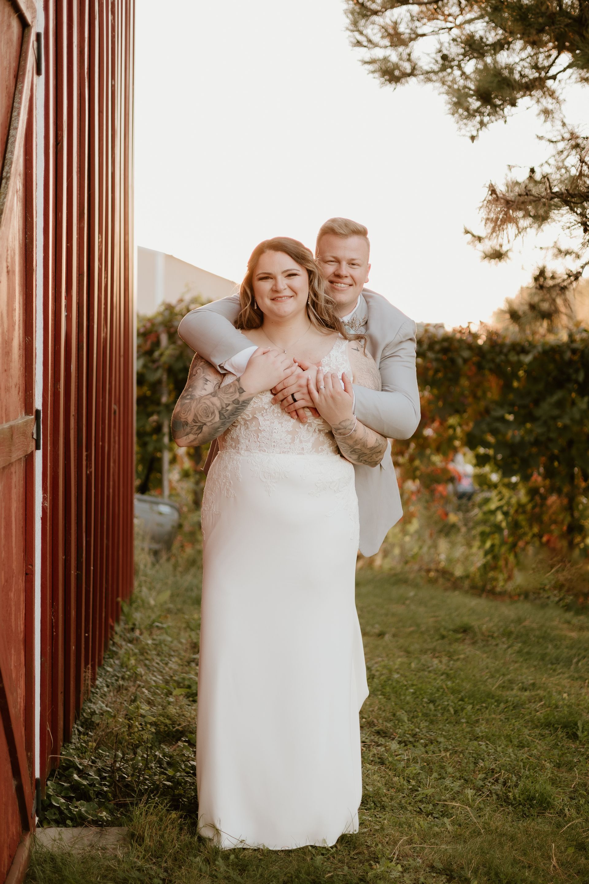 Two people in wedding attire standing outdoors by a red wooden wall, smiling and holding bouquets