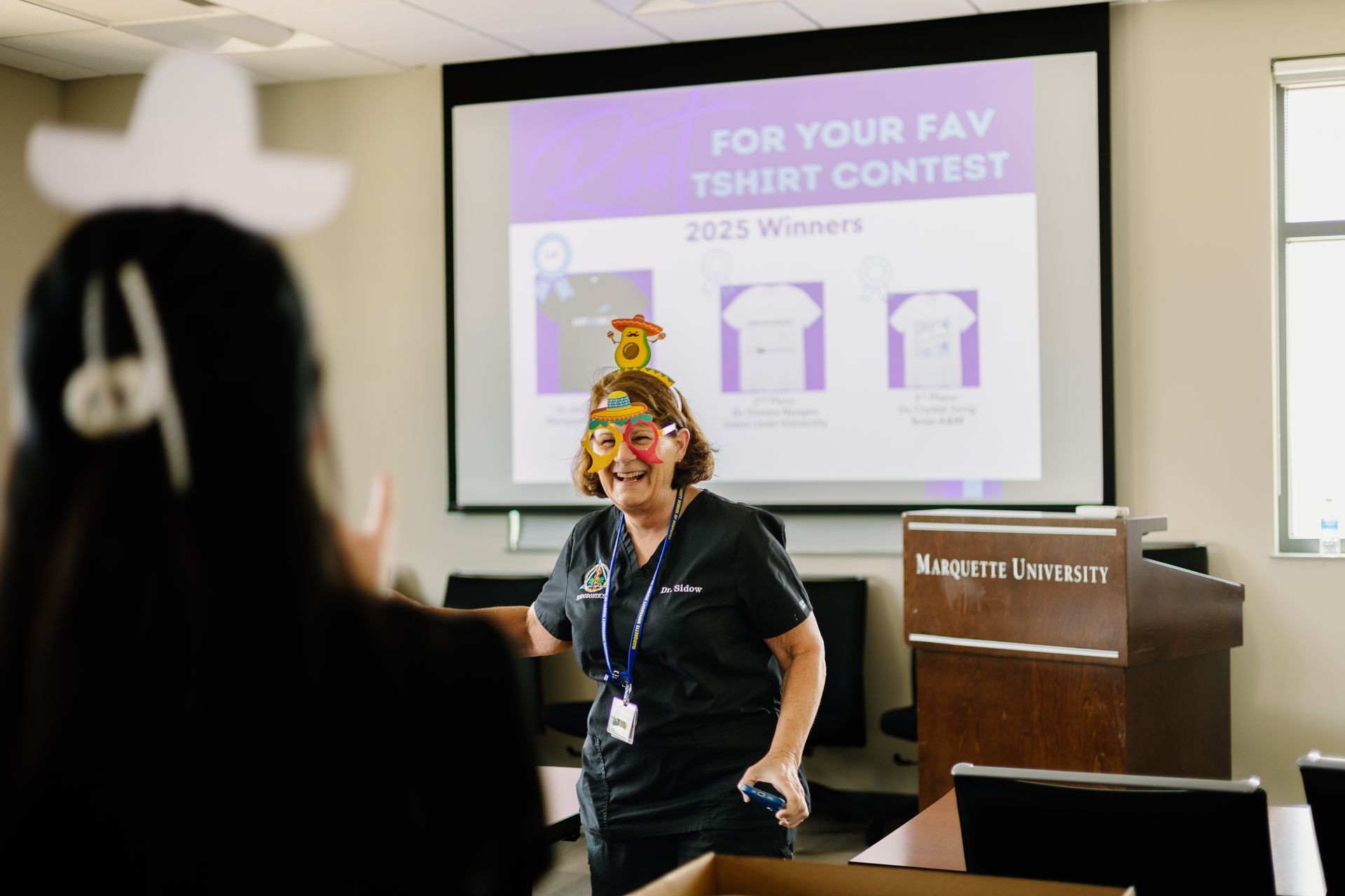 Woman presenting in a classroom beside a slide about a T-shirt contest
