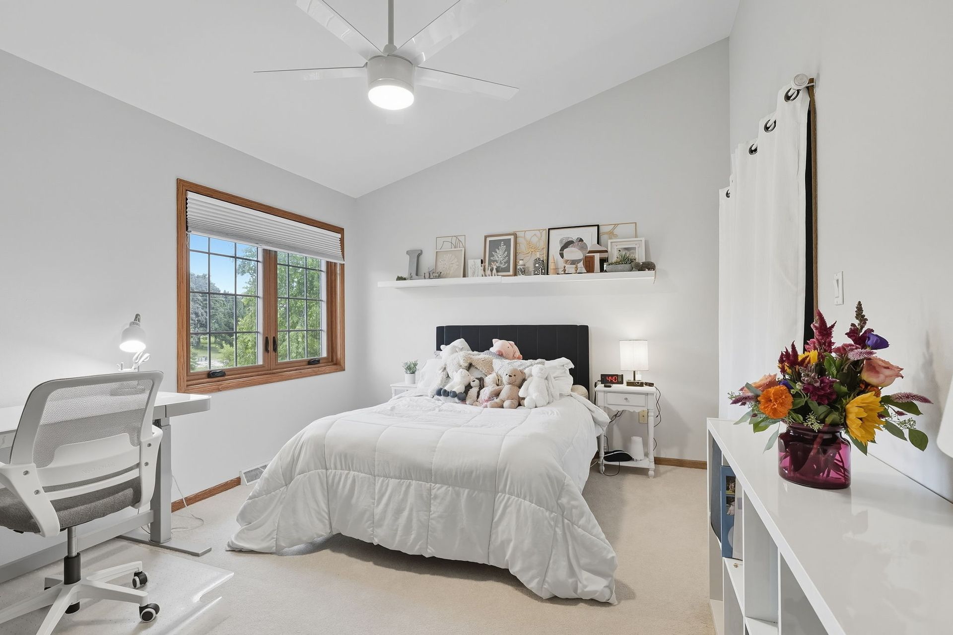 Bright white bedroom with a bed, ceiling fan, window, and a colorful bouquet on a dresser.