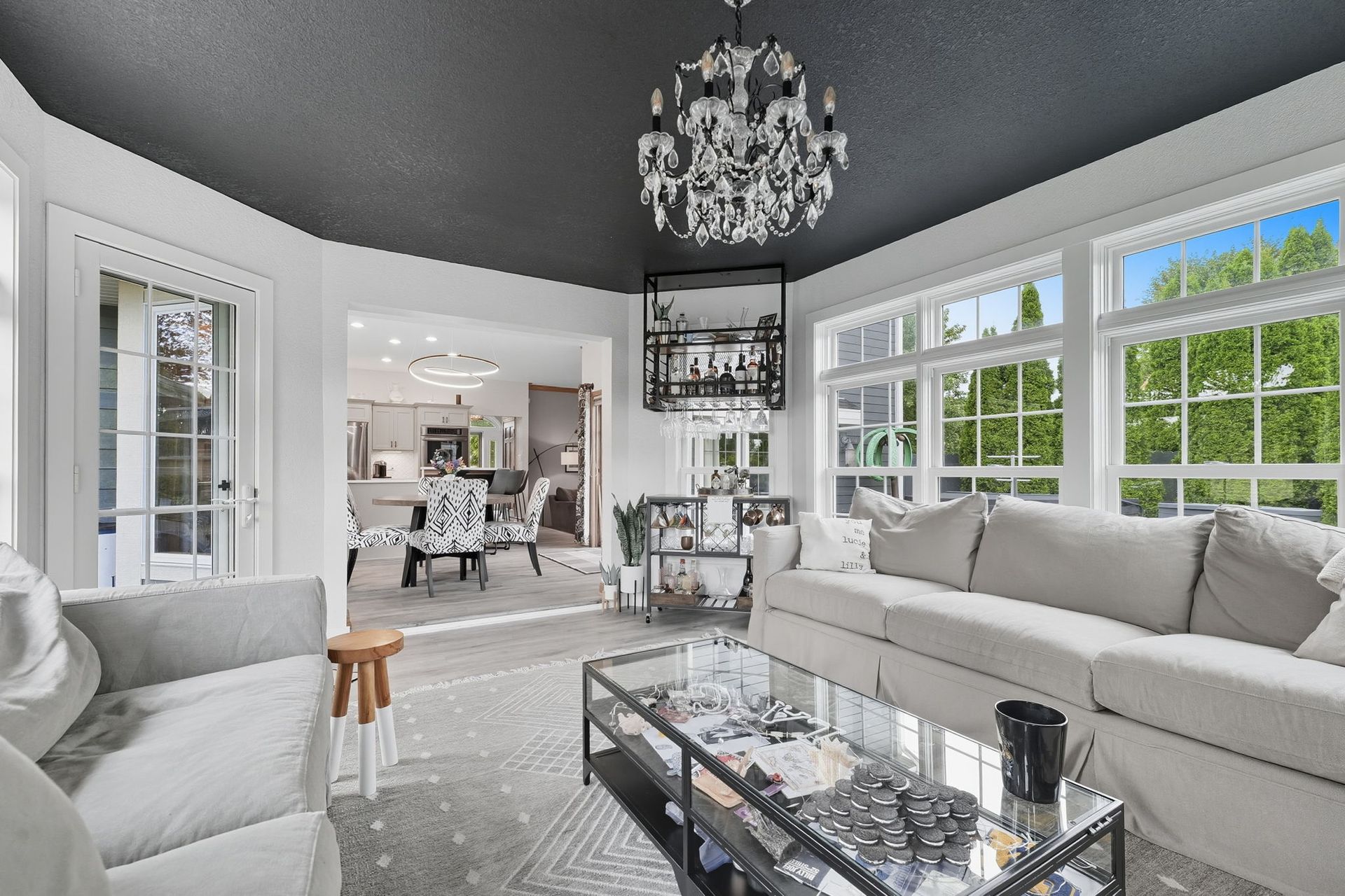 Bright modern living room with white sofas, glass coffee table, chandelier, and large windows.