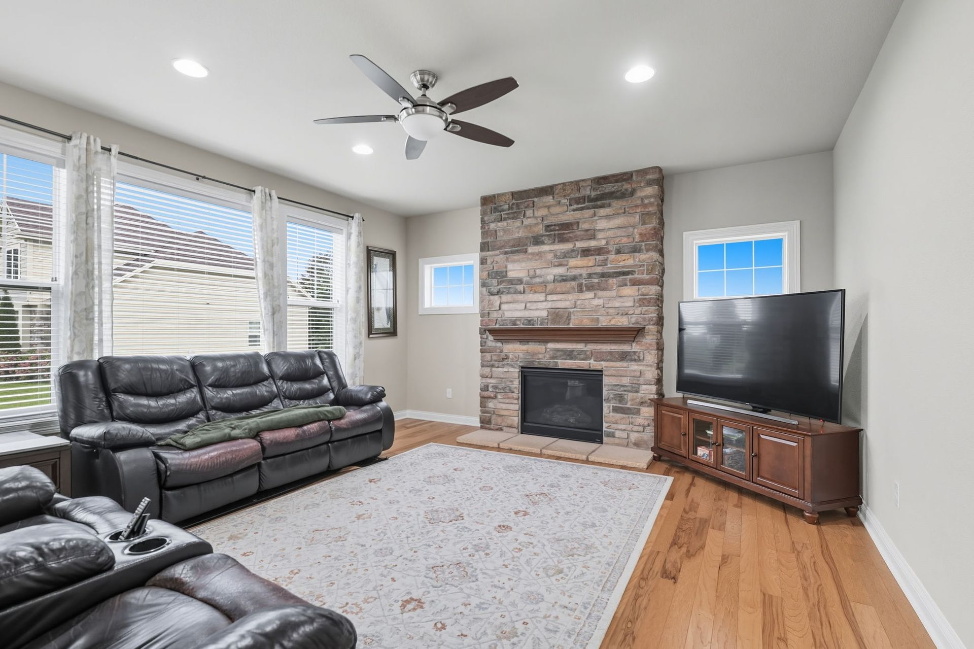 Living room with leather sofas, large windows, brick fireplace, TV, and ceiling fan