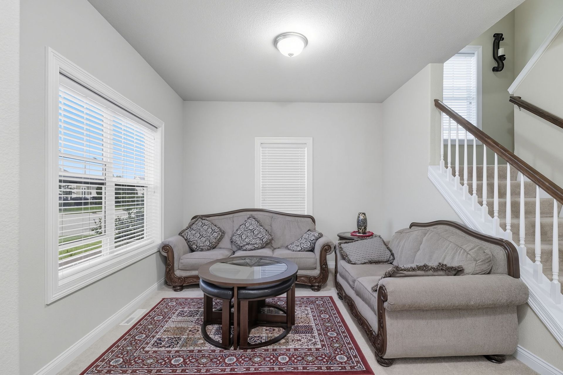 Bright sitting room with two gray sofas, patterned rug, coffee table, and staircase beside a window.