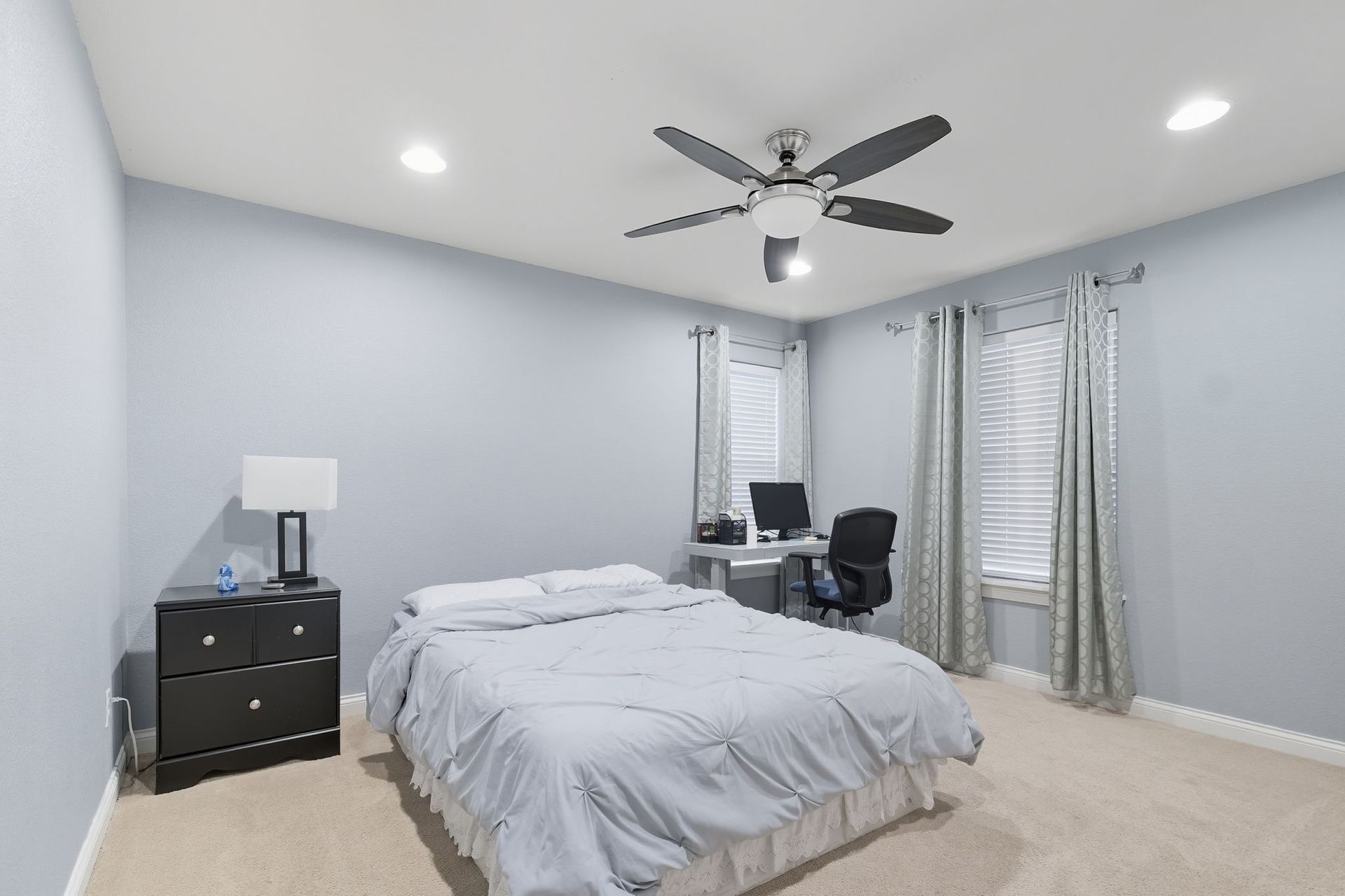 Bright bedroom with a made bed, black dresser, desk, and large windows with curtains