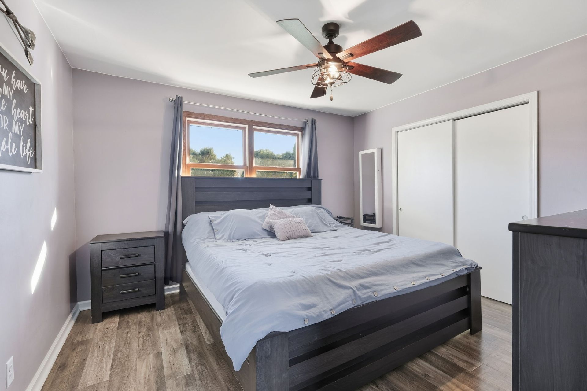 Bright bedroom with a bed, nightstand, ceiling fan, and large window with curtains